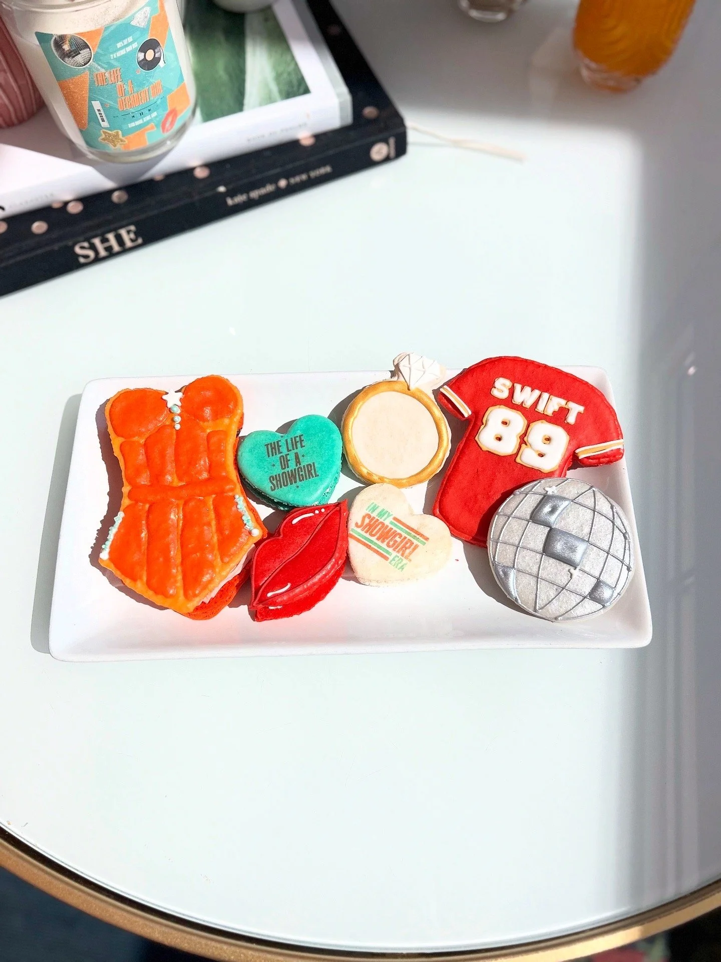 A white rectangular plate with four decorative cookies and two cupcakes, placed on a white table. The cookies are shaped like a dress, a volleyball, lips, and a heart with the words "The Life of a Showgirl" and "In My Showgirl Era." The cupcakes are decorated with a red sports jersey and a silver volleyball, surrounded by other colorful items, with a partially visible book or magazine and drinks in the background.