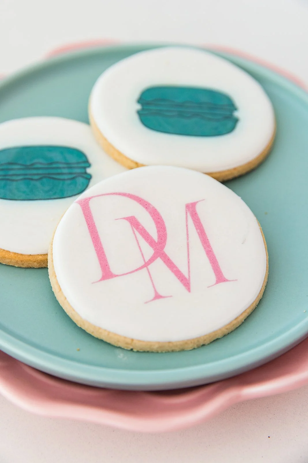Decorated cookies on a pastel dish, featuring a pink monogram and a blue and white design resembling a dessert or macaron — custom dessert catering and events by Decadent Macaron in Gilbert, Arizona