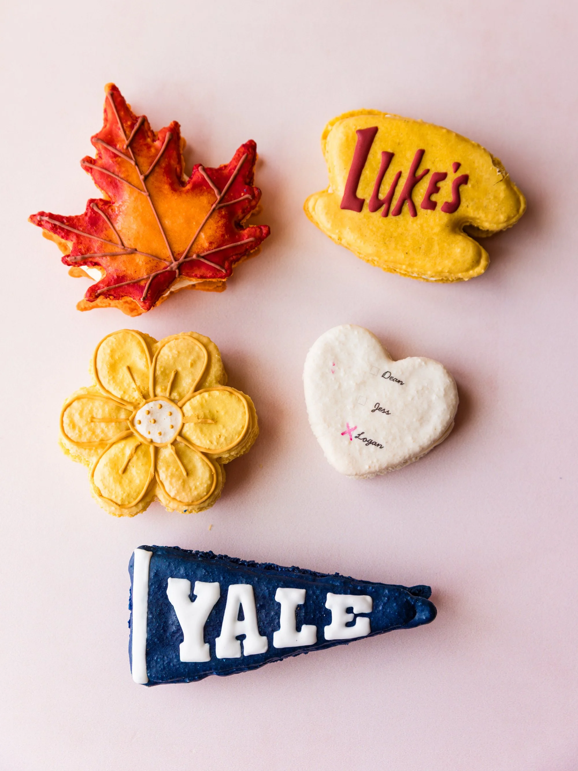 Cookies decorated with fall and school themes, including a red maple leaf, a yellow flower, a yellow cloud with LUKES written on it, a heart-shaped cookie with handwritten names, and a blue YALE pennant — custom dessert catering and events