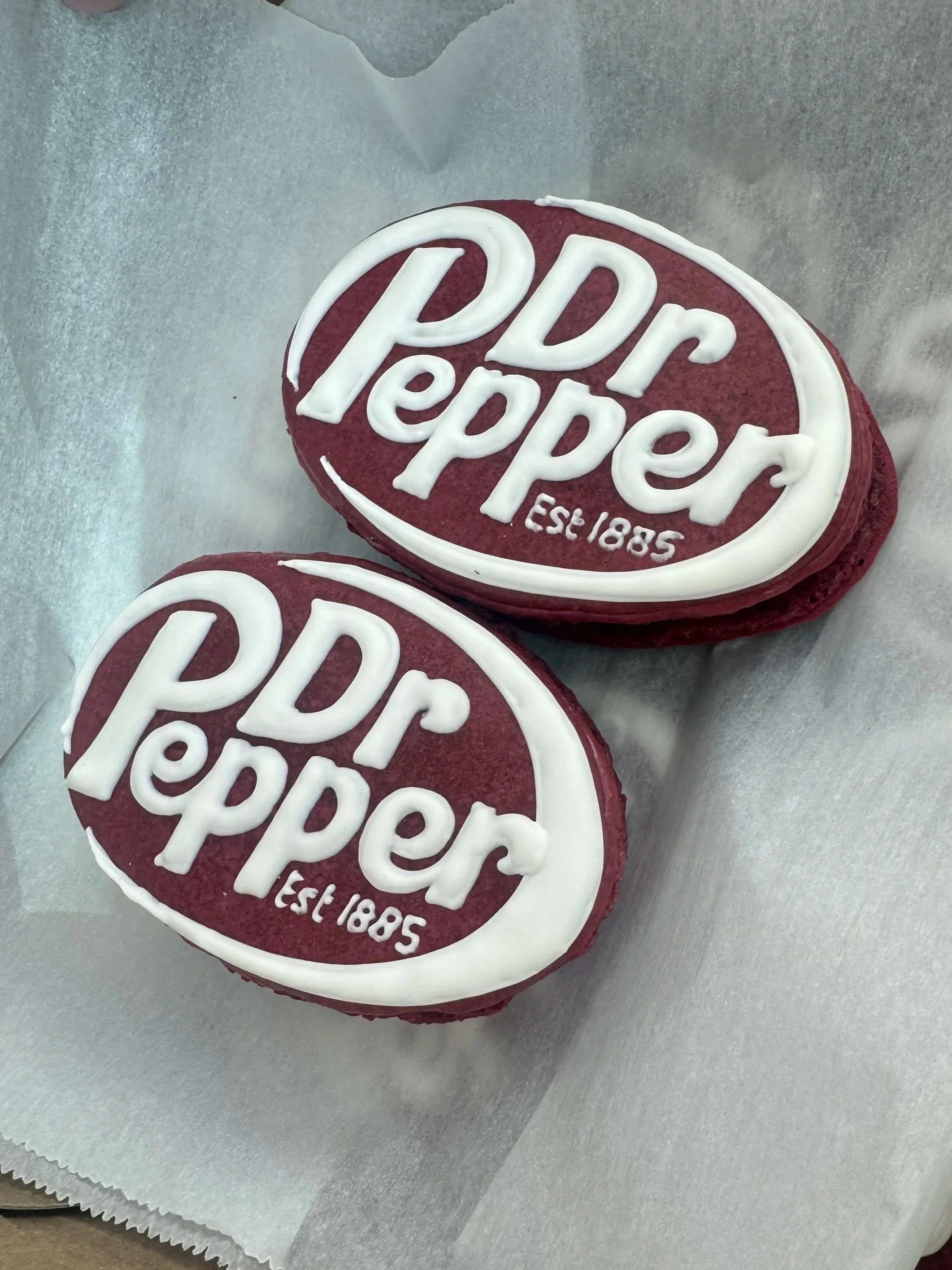 Two oval cookies with red icing and white lettering that says "Dr Pepper" and "Est. 1885" resting on parchment paper — custom dessert catering and events by Decadent Macaron in Gilbert, Arizona
