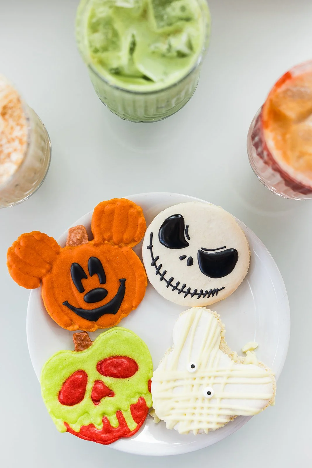 Halloween-themed cookies with orange Mickey Mouse shape, white skull face, green and red skull, and a white ghost, along with drinks in glasses around a white plate — handcrafted macarons and desserts at Decadent Macaron café in Gilbert, Arizona