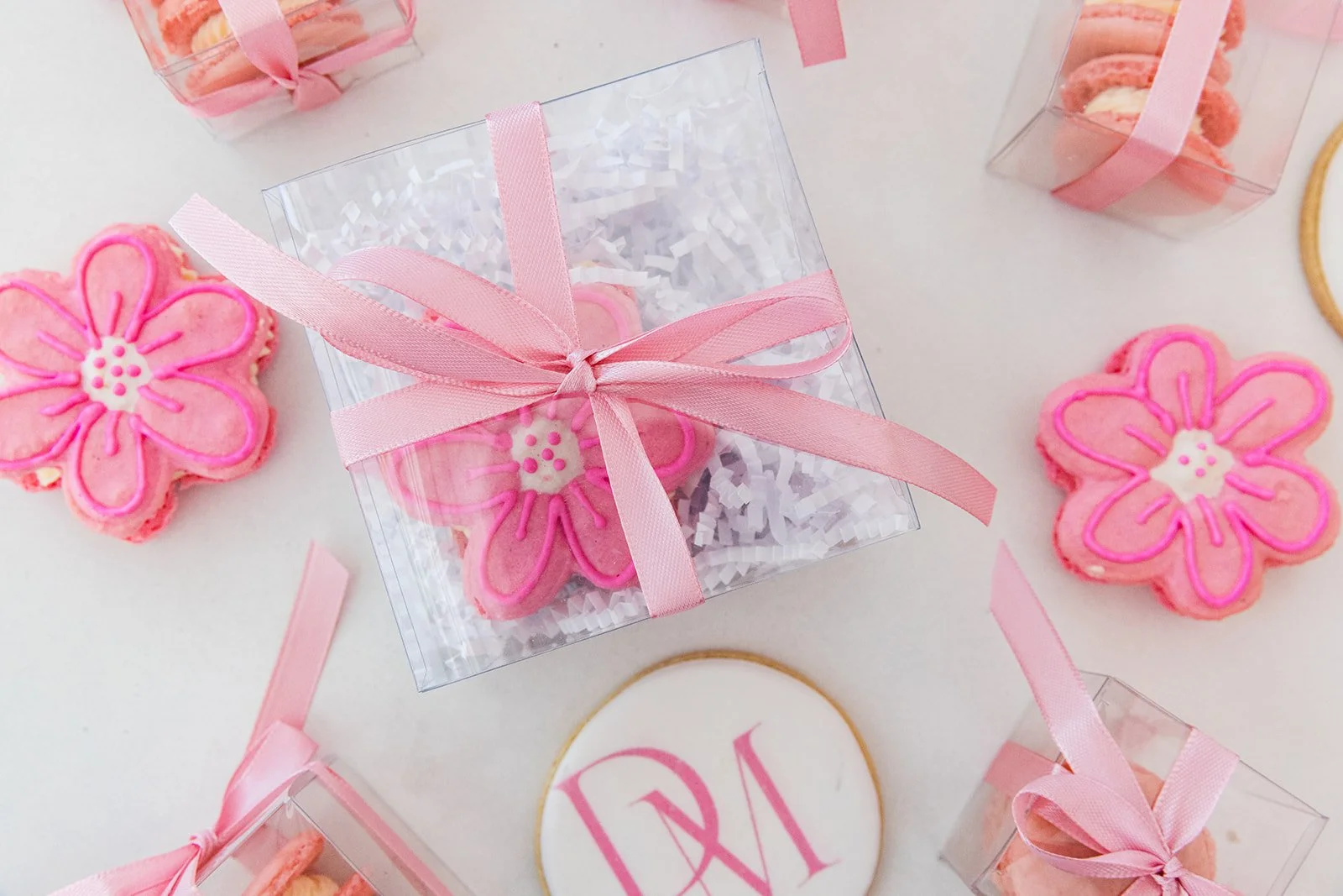 Pink flower-shaped cookies decorated with pink icing, some wrapped in transparent boxes with pink ribbons, on a white surface — custom dessert catering and events by Decadent Macaron in Gilbert, Arizona
