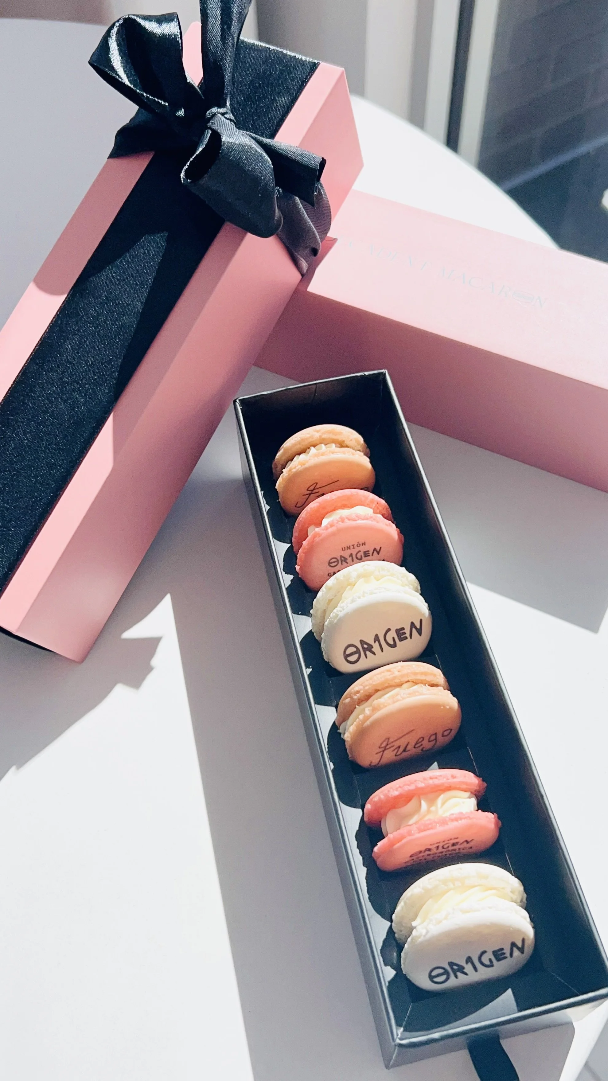 A box of colorful macarons on a white surface, with a pink gift box featuring a black ribbon next to it — custom dessert catering and events by Decadent Macaron in Gilbert, Arizona