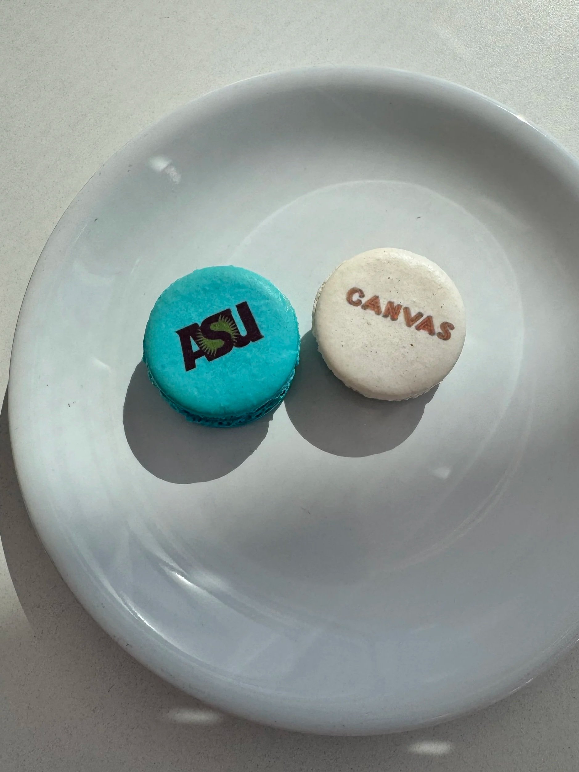 Two macarons on a white plate, one blue with the word 'ASIL' and one white with the word 'CANVAS' written on them — custom dessert catering and events by Decadent Macaron in Gilbert, Arizona