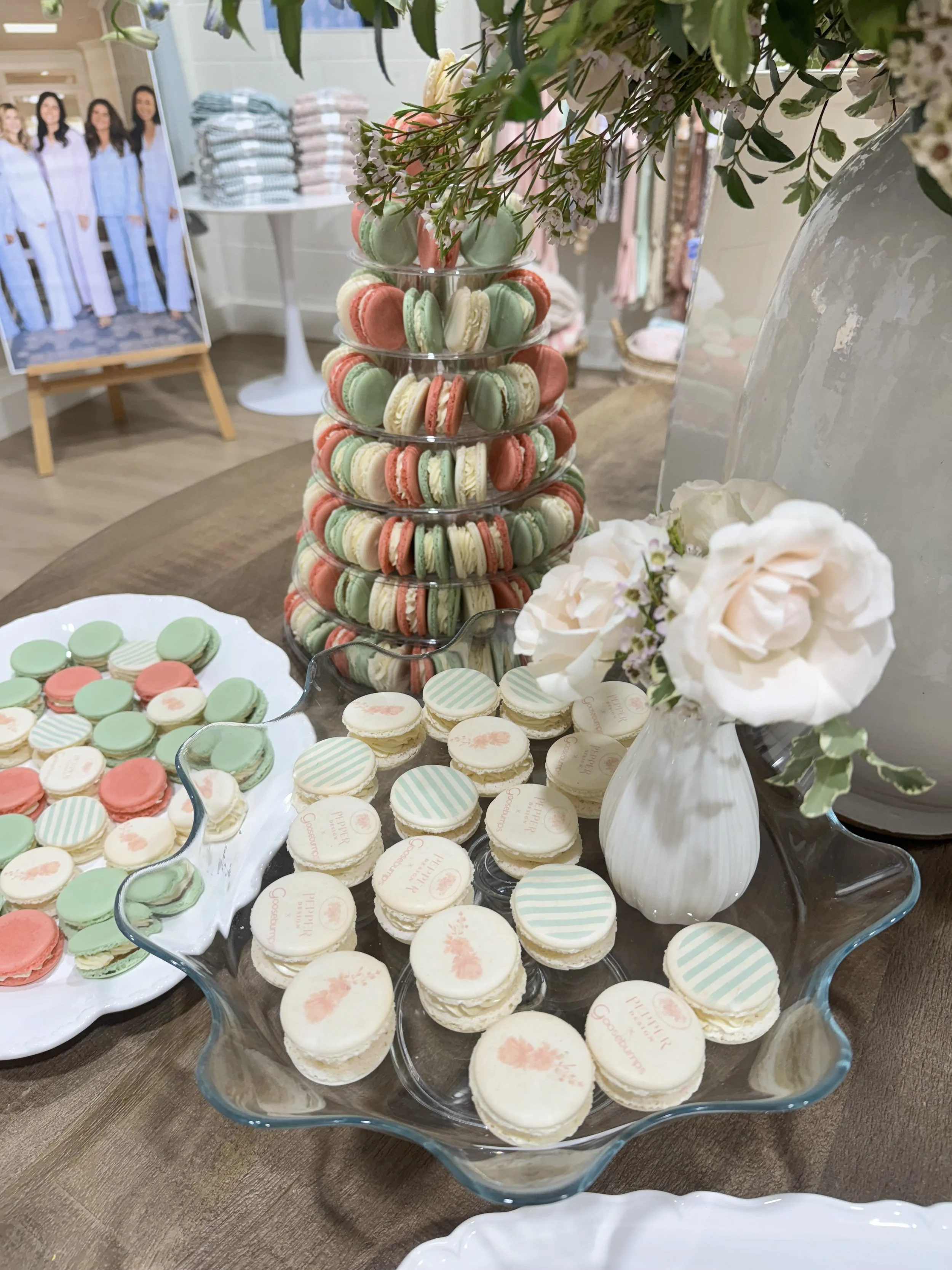 Display of pastel-colored macarons and cookies on glass trays, with a floral arrangement in a white vase on a wooden table — custom dessert catering and events by Decadent Macaron in Gilbert, Arizona