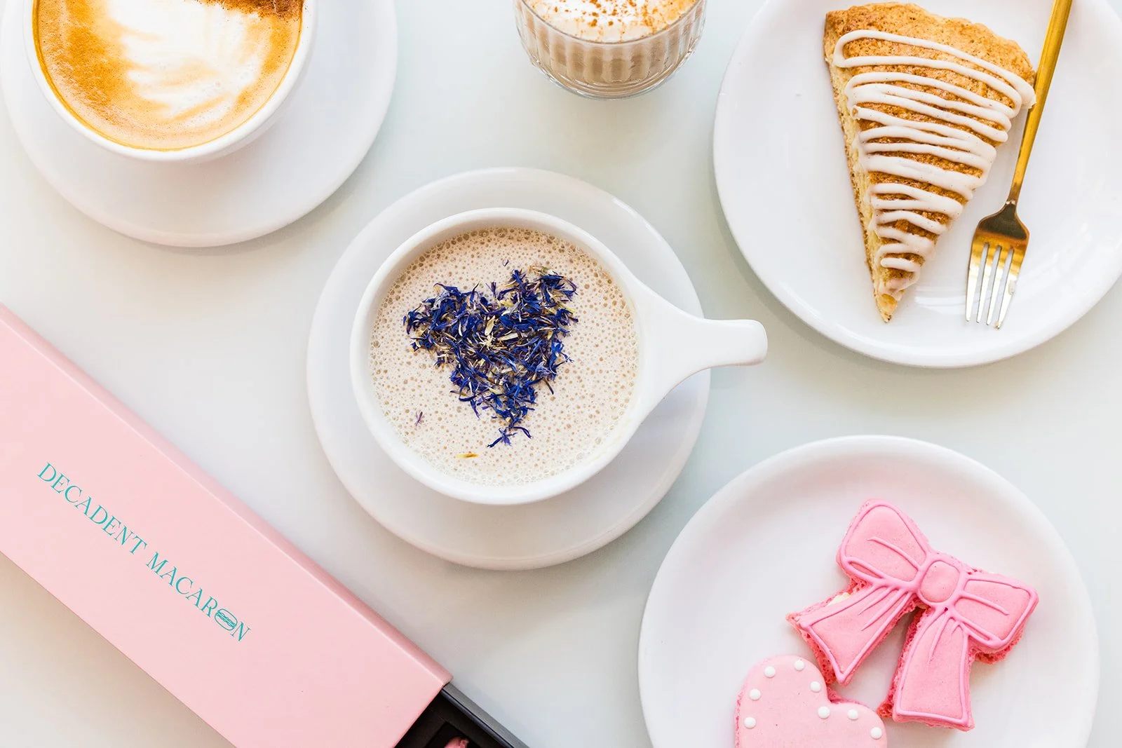 A breakfast spread including a cup of coffee with foam, a glass of a creamy drink, a plate with a slice of lemon cake with icing, a mug of coffee with purple flower petals heart-shaped on top, a plate with pink decorated cookies, and a pink box labeled "Decadent Macaron".