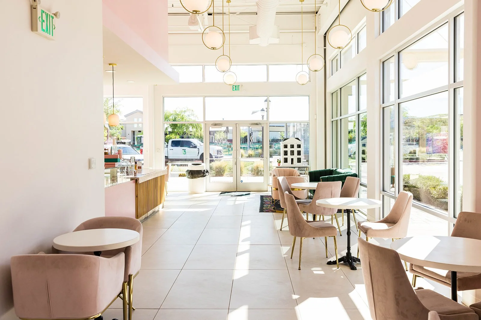 Bright interior of a modern café with pink velvet chairs, white round tables, large windows, and pendant lights, with the entrance and outdoor scene visible — custom dessert catering and events by Decadent Macaron in Gilbert, Arizona