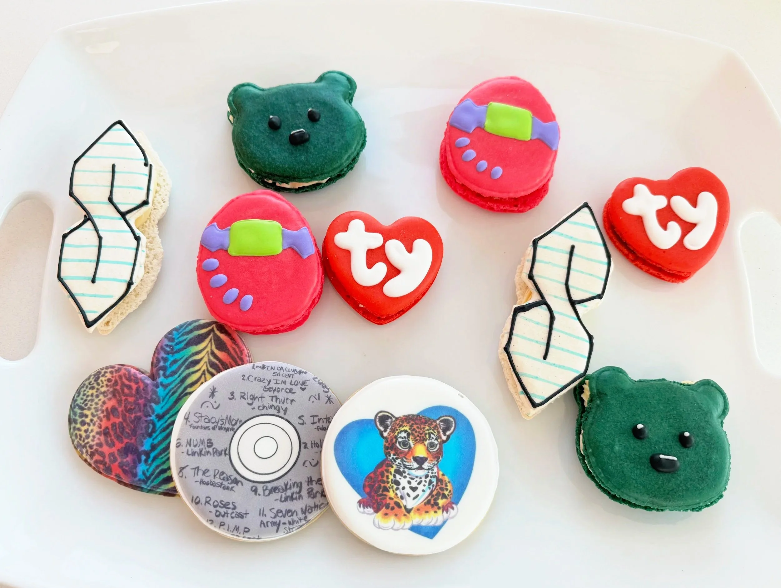 Assorted colorful cookies on a white tray, including bear-shaped cookies, heart-shaped cookies with 'ty' and 'try' messages, ice-cream cone-shaped cookies, and a cookie with a leopard illustration and a handwritten song list — custom dessert catering