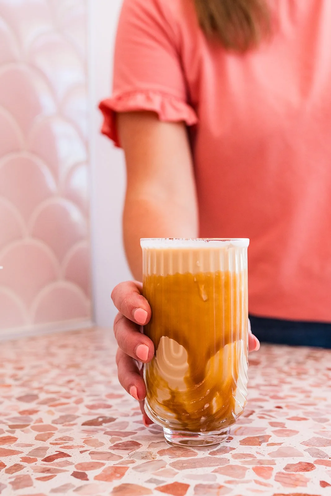 A person in a pink shirt holding a glass of iced coffee or latte on a speckled pink and white counter with a pink scalloped wall in the background — Decadent Macaron café in Gilbert, Arizona dessert shop and specialty café