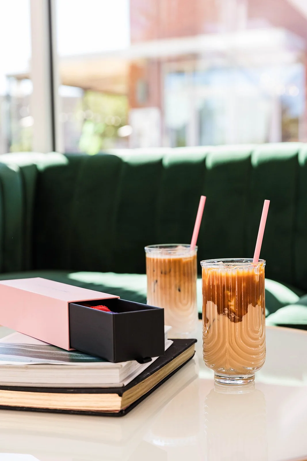 Two glasses of iced coffee with pink straws on a white table, next to a pink box and books, in a bright café with green seating and large windows — Decadent Macaron café in Gilbert, Arizona dessert shop and specialty café