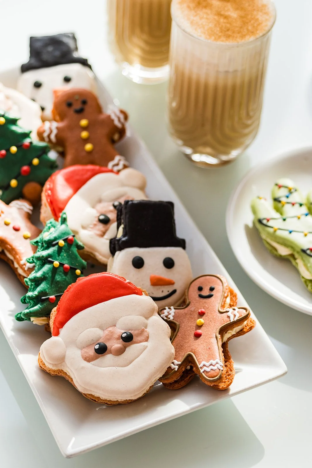 Assorted Christmas cookies shaped like Santa Claus, snowmen, gingerbread men, and Christmas trees, with two glasses of iced coffee and a plate of decorated green cookies in the background — handcrafted macarons and desserts at Decadent Macaron café