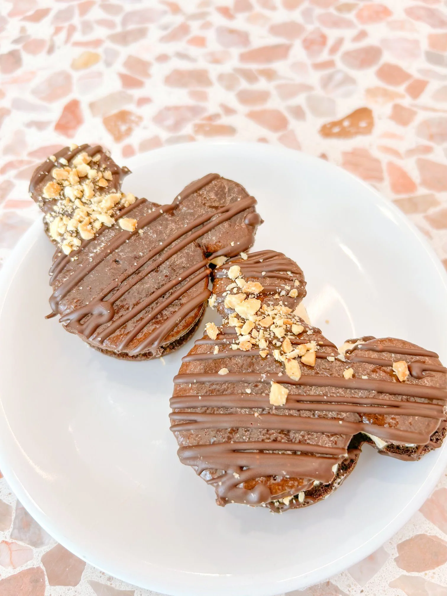 Two chocolate-covered, heart-shaped cookies with slanted chocolate drizzle and sprinkled chopped nuts on top, placed on a white plate — handcrafted macarons and desserts at Decadent Macaron café in Gilbert, Arizona