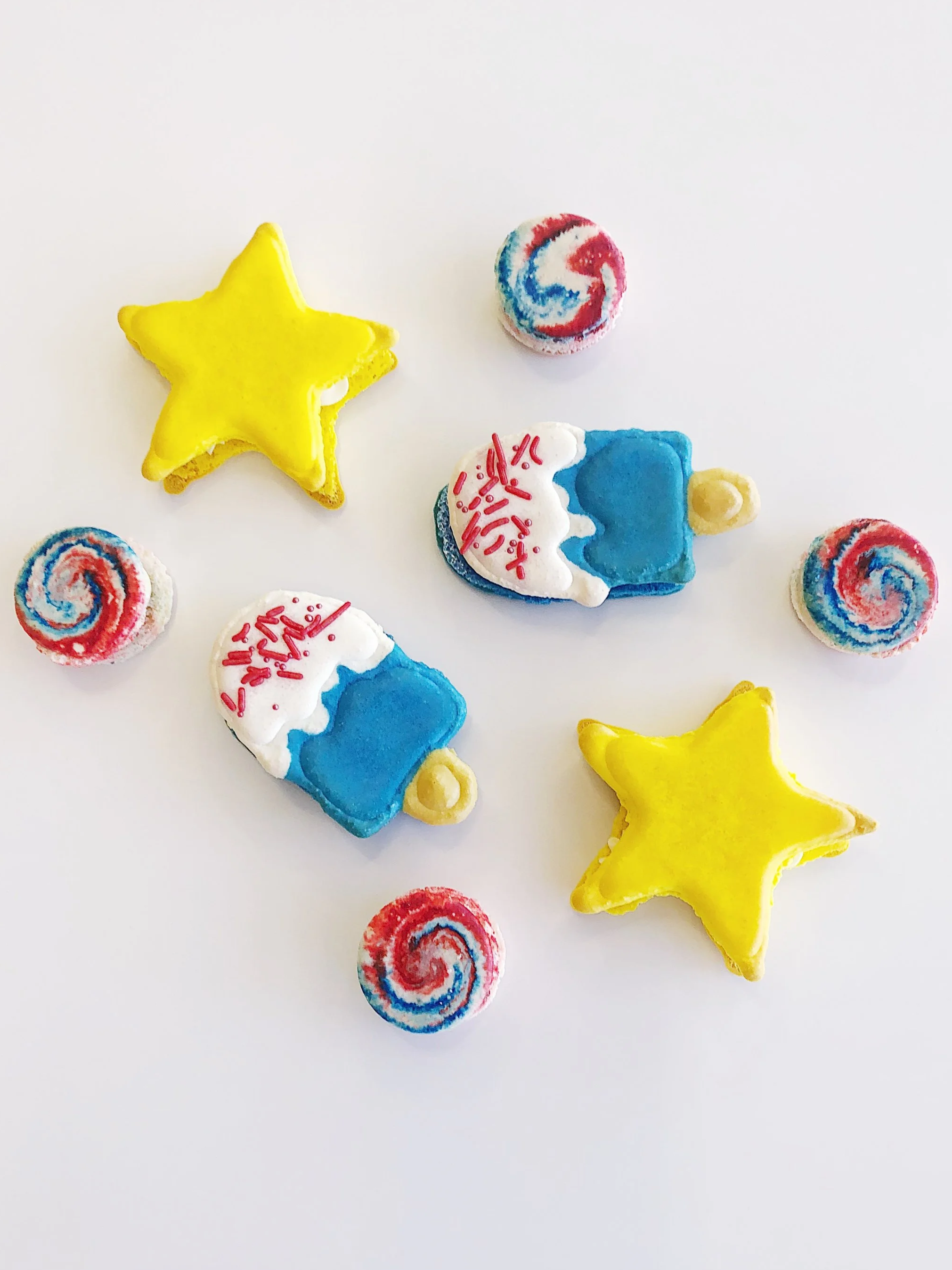 Colorful decorative candies shaped like yellow stars, red and blue swirled lollipops, and ice cream pops with blue, white, and red icing and sprinkles on a white background — custom dessert catering and events by Decadent Macaron in Gilbert, Arizona