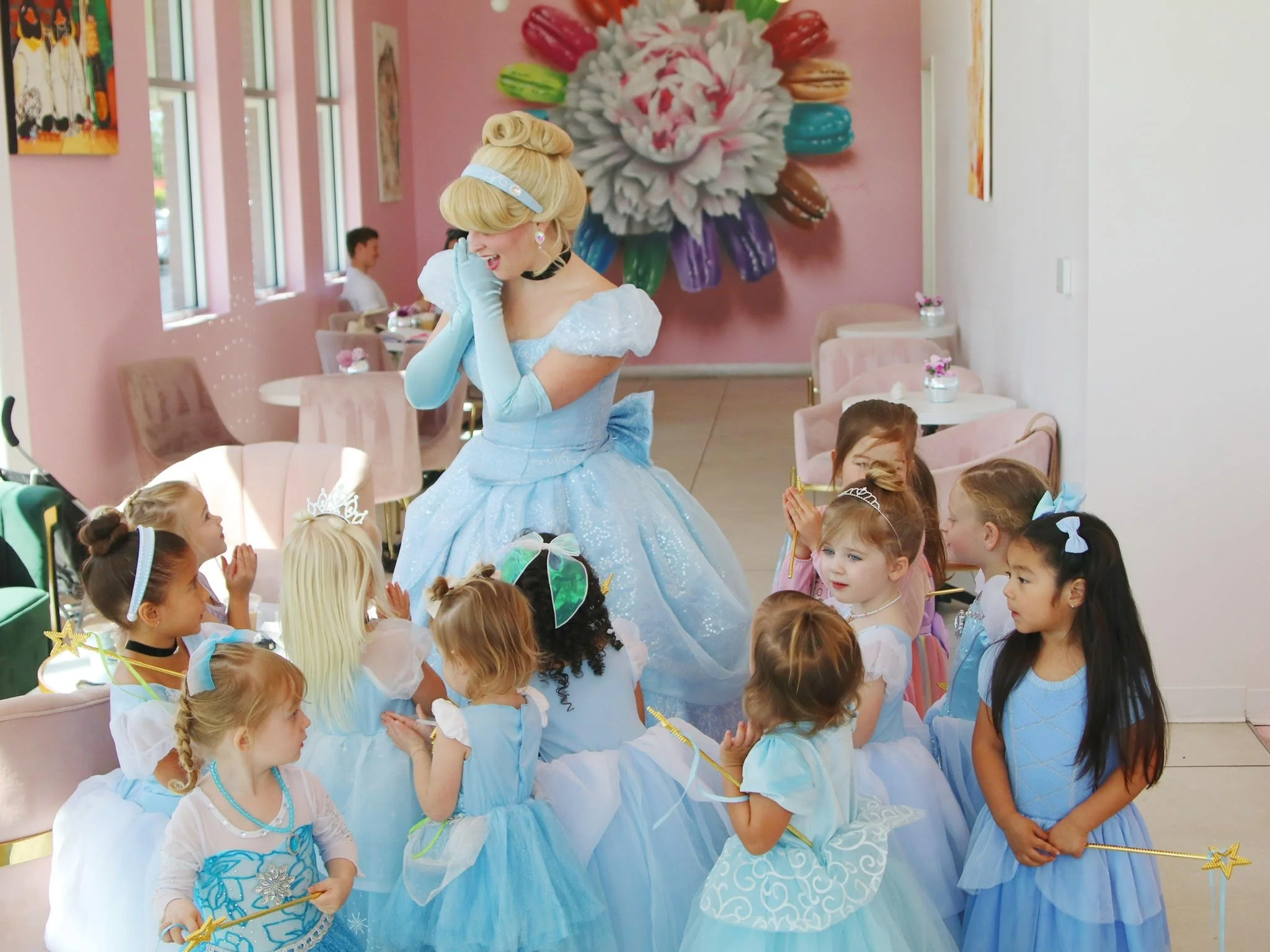 A woman dressed as Cinderella surrounded by young girls in princess dresses at a tea party in a decorated pink room — custom dessert catering and events by Decadent Macaron in Gilbert, Arizona