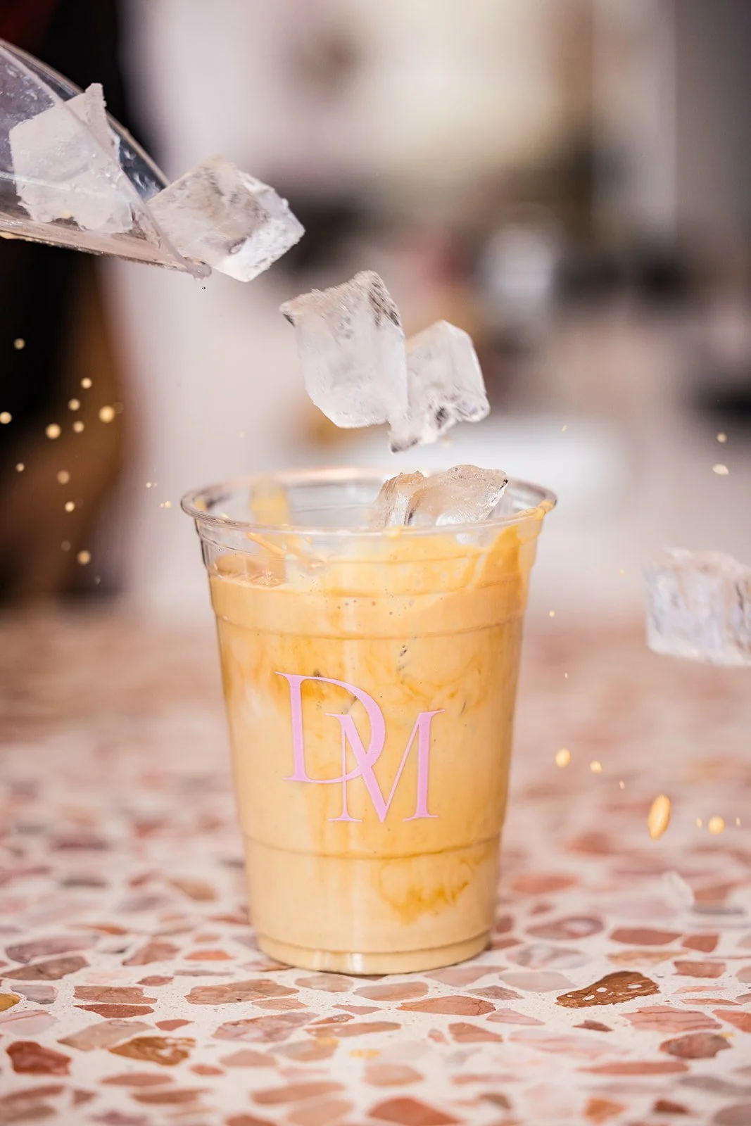 A creamy, orange-colored beverage in a clear plastic cup with a pink monogram logo, ice cubes being added on top, on a tiled surface — Decadent Macaron café in Gilbert, Arizona dessert shop and specialty café