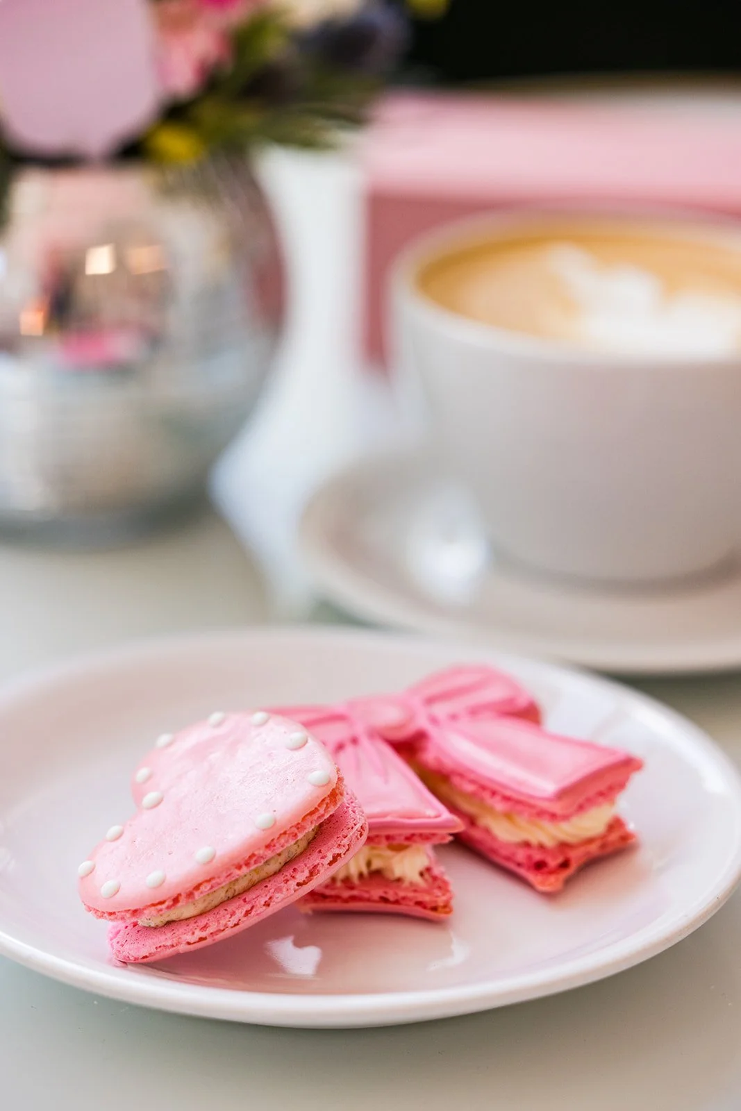 Pink heart-shaped macarons with white filling on a white plate, a cup of coffee with latte art in the background, and a flower arrangement in a reflective vase — handcrafted macarons and desserts at Decadent Macaron café in Gilbert, Arizona