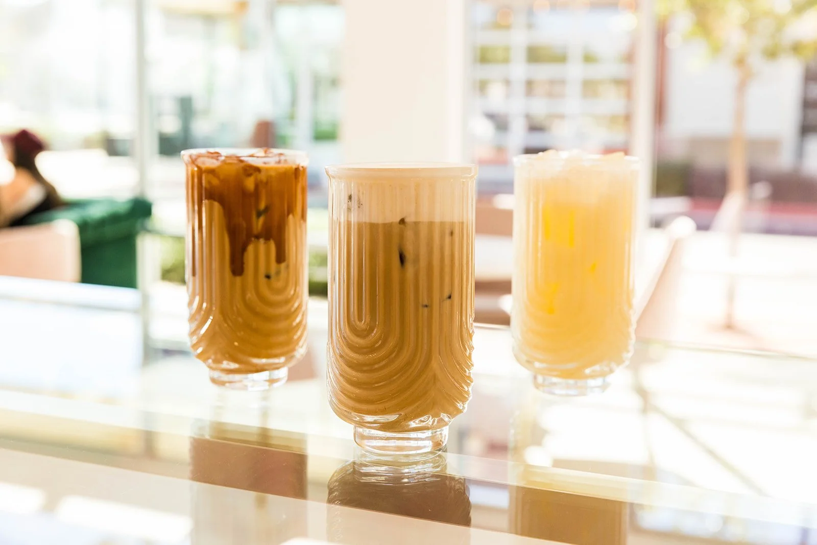 Three tall glasses with iced coffee drinks, each with different shades of brown and yellow, placed on a shiny white table in a bright indoor setting — Decadent Macaron café in Gilbert, Arizona dessert shop and specialty café