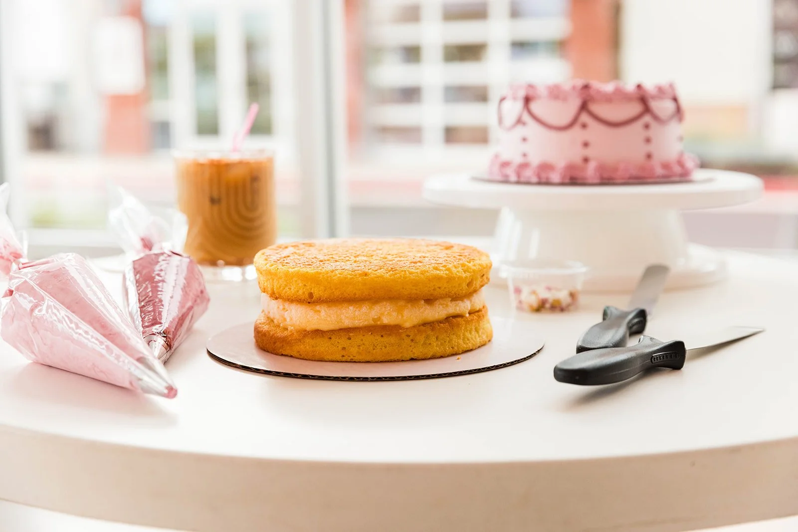 A birthday cake with pink icing and decorative piping on a cake stand in the background, a partially assembled ice cream sandwich with cookies and ice cream in the foreground, a pink star-shaped piping bag, a pair of black scissors, and a glass of iced coffee on a white table.