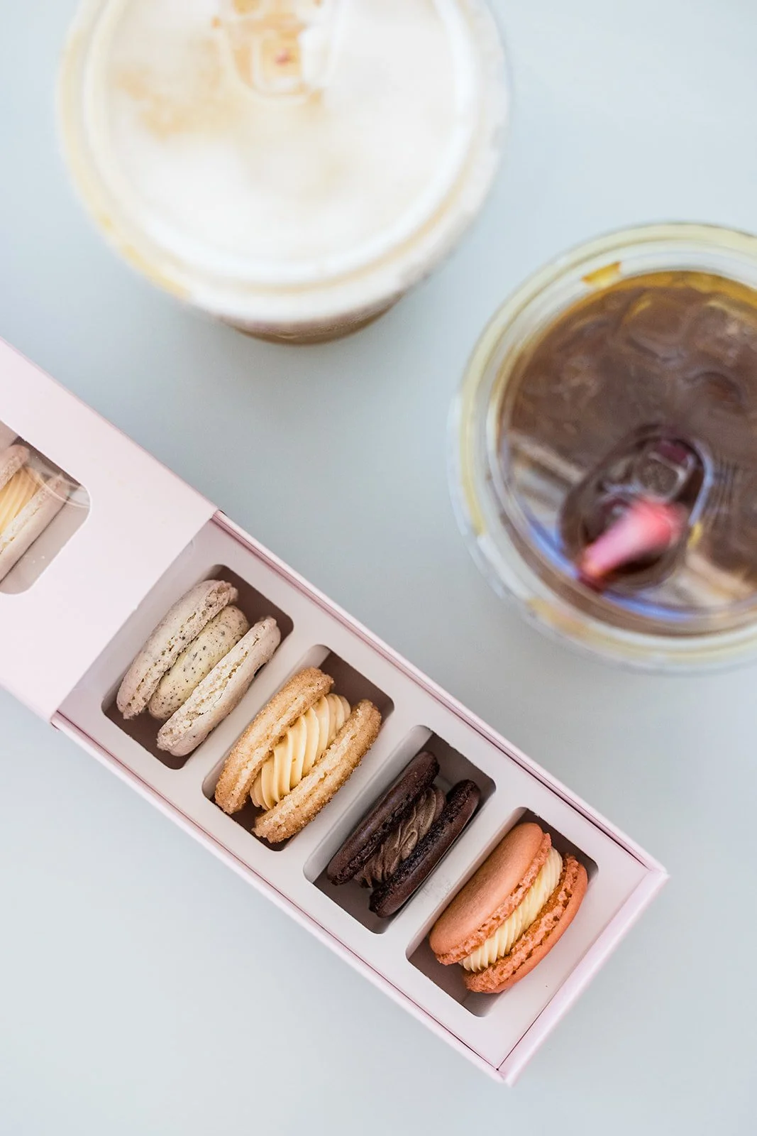 A box of assorted macarons on a light-colored table with two iced coffee drinks in cute cups nearby — handcrafted macarons and desserts at Decadent Macaron café in Gilbert, Arizona