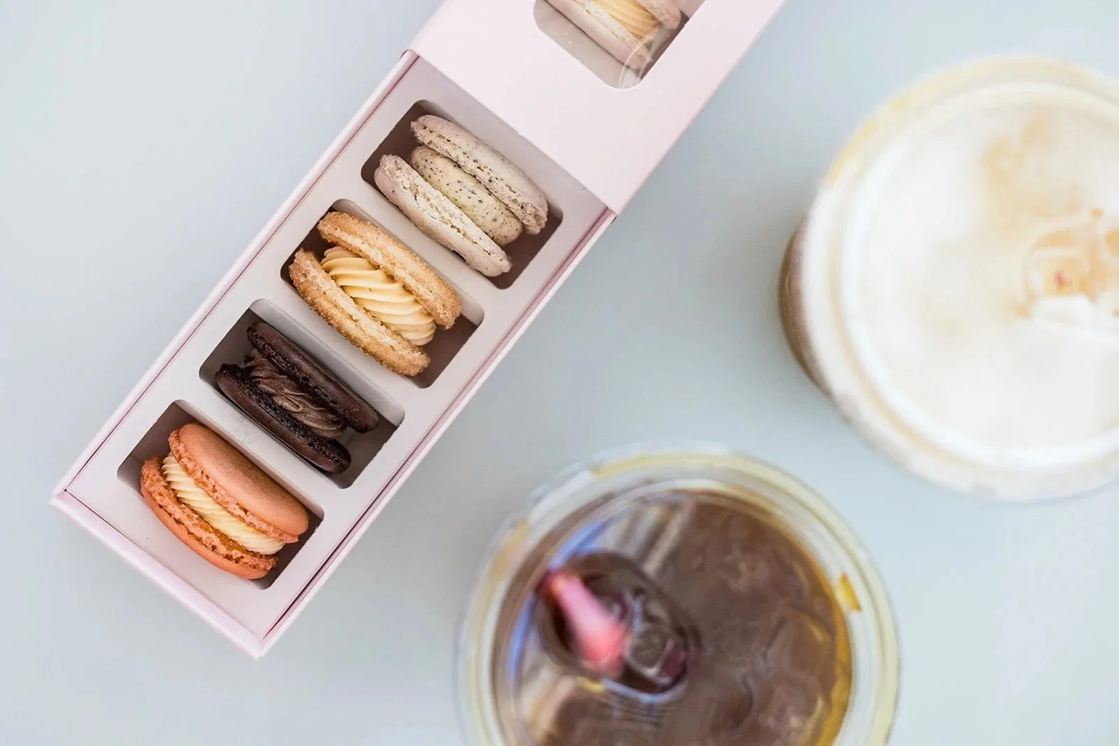 A box of assorted macarons next to two beverages, one dark and one light, on a light surface — Decadent Macaron café in Gilbert, Arizona dessert shop and specialty café