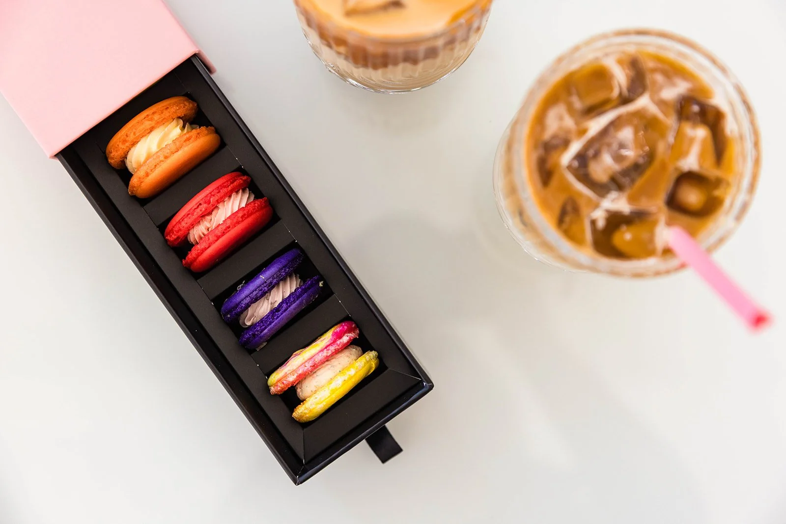 Colorful macarons in a black box with a pink top, and two glasses of iced coffee with ice cubes and straws on a white surface — handcrafted macarons and desserts at Decadent Macaron café in Gilbert, Arizona