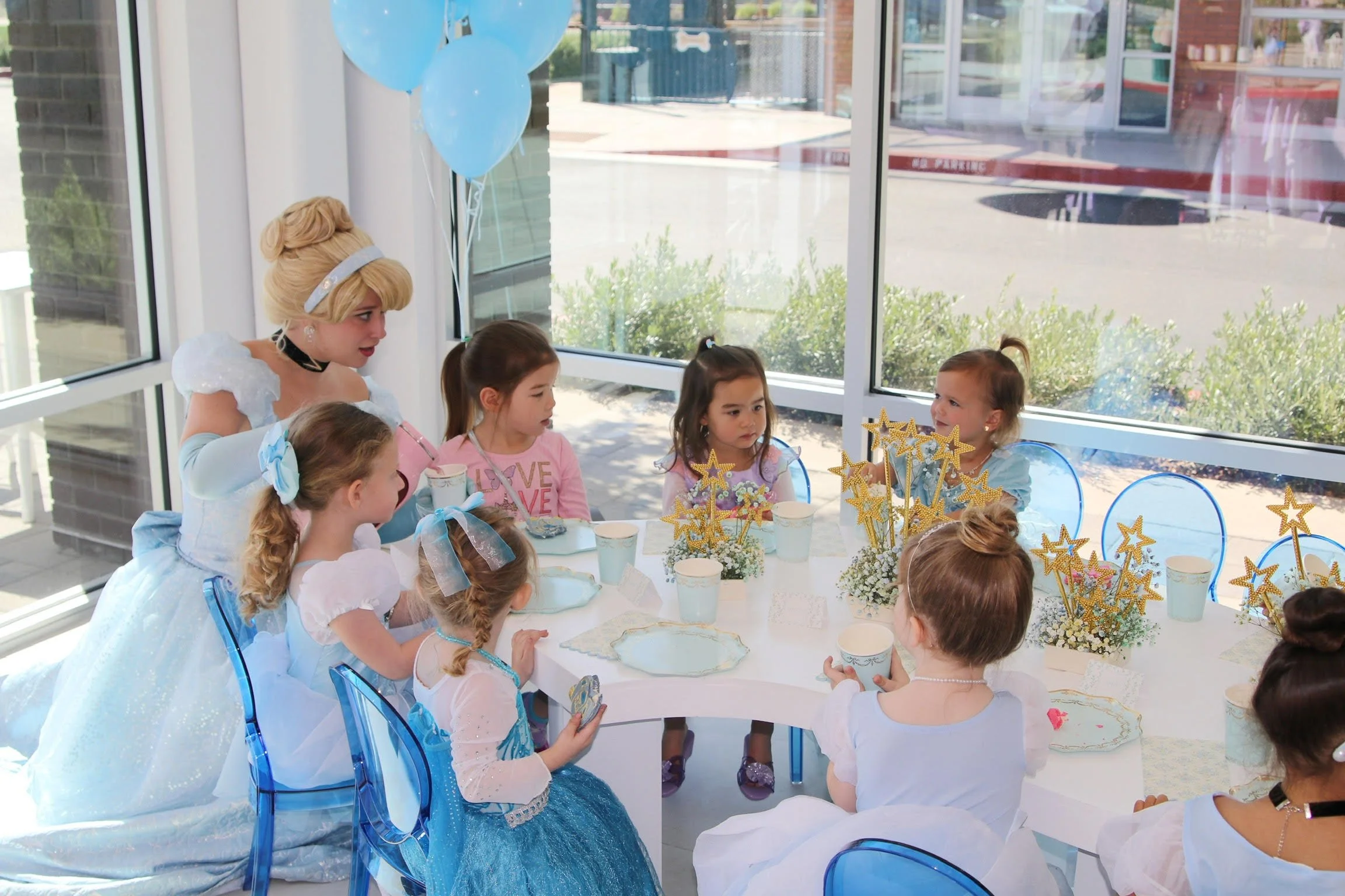 A princess dressed in a light blue gown and tiara with blonde hair, supervising a girl in a princess dress holding a plate at a children's birthday party with fairy-tale theme, including balloons, star-shaped wands, and decorated table — custom desse