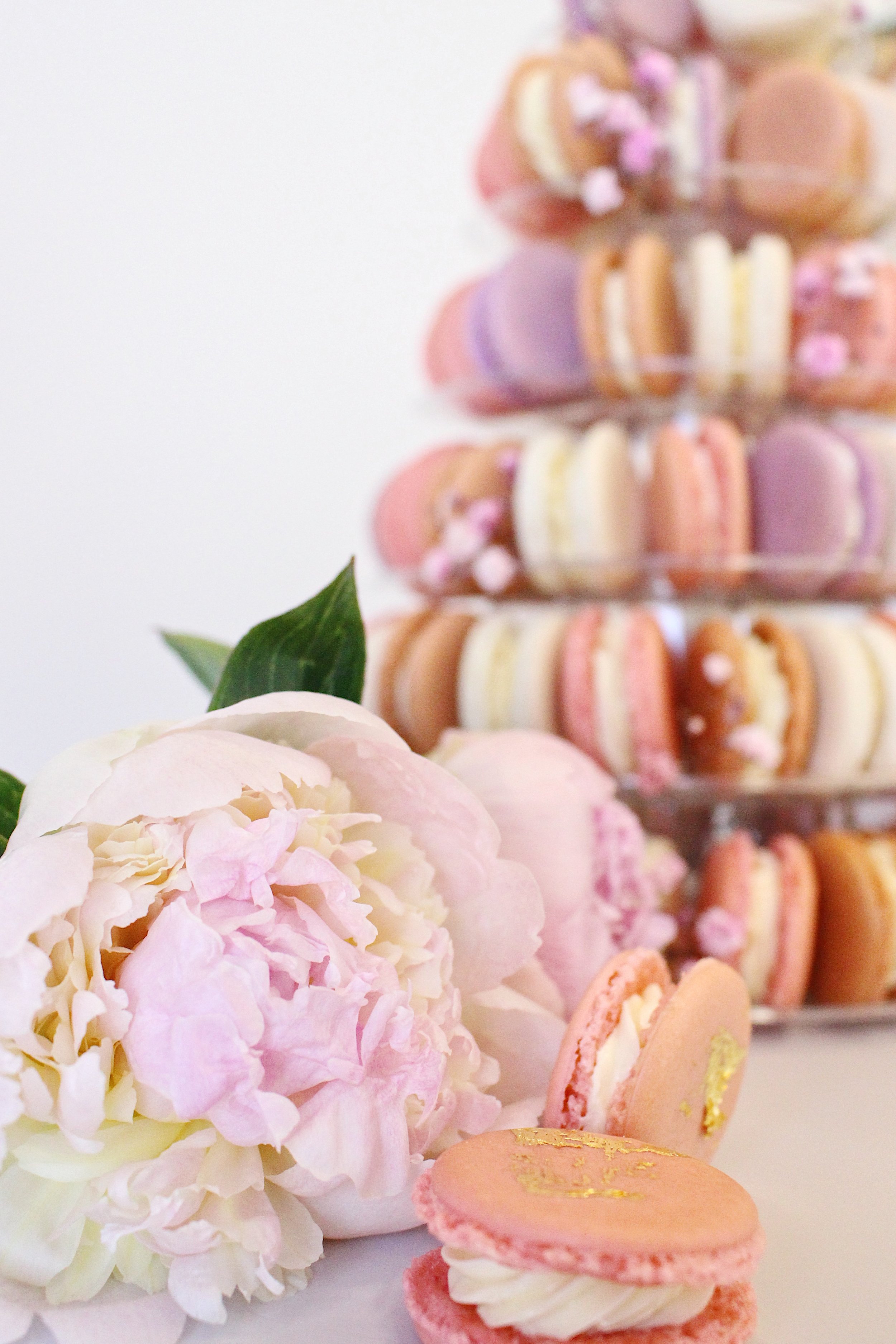 Pink and white peonies with pastel-colored macarons in front of a tiered display of macarons — custom dessert catering and events by Decadent Macaron in Gilbert, Arizona
