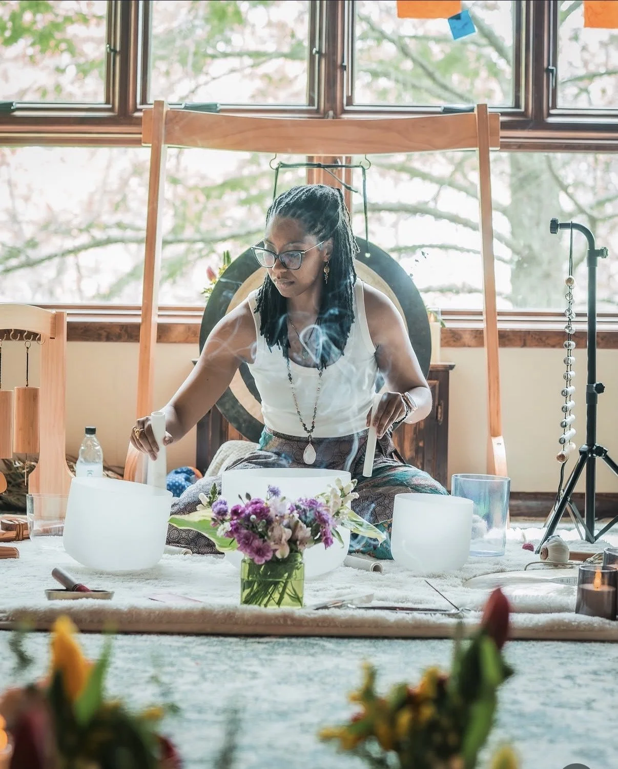 A woman with glasses and dreadlocks is sitting on the floor, playing crystal singing bowls. She is surrounded by flowers, candles, and other musical instruments in a bright room with large windows.