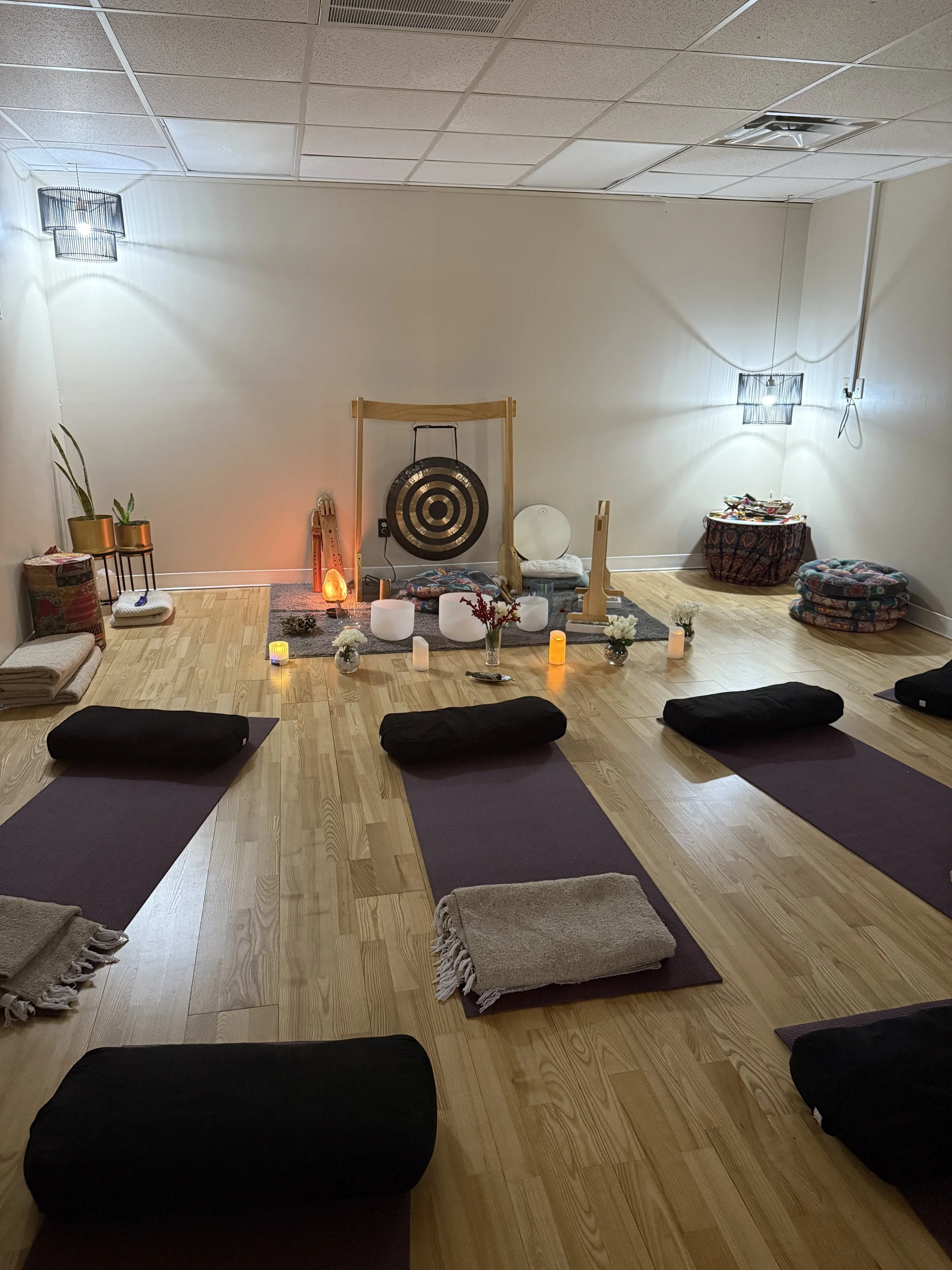 A yoga or meditation space with mats and pillows arranged on a wooden floor. Candles, flowers, and a salt lamp are placed on a rug in the center, creating a calming atmosphere. The room has white walls and ceiling lights with decorative fixtures.
