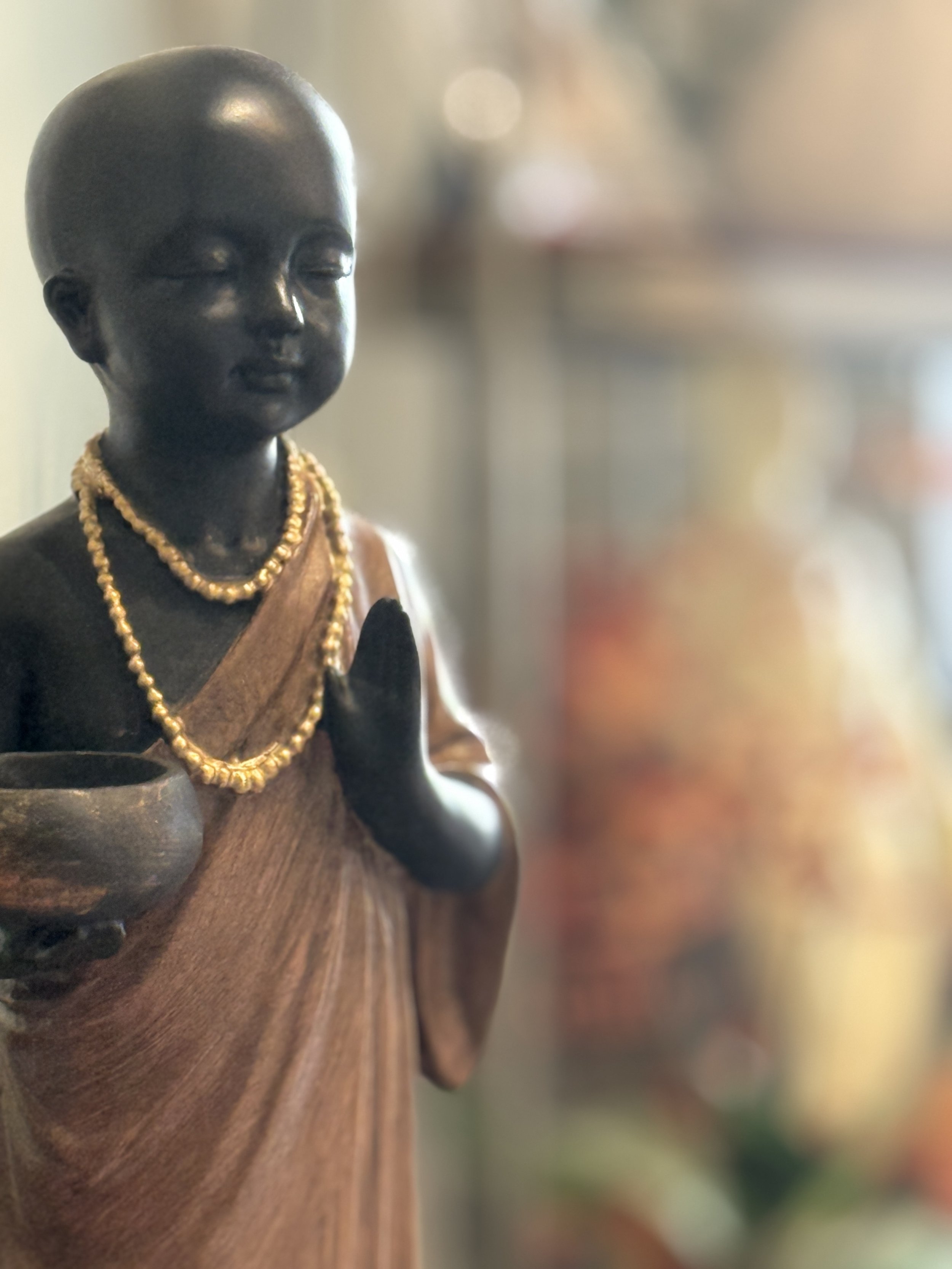 A close-up of a dark figurine of a child or deity with closed eyes, wearing gold jewelry, mounted on a wooden surface with a blurred colorful background.