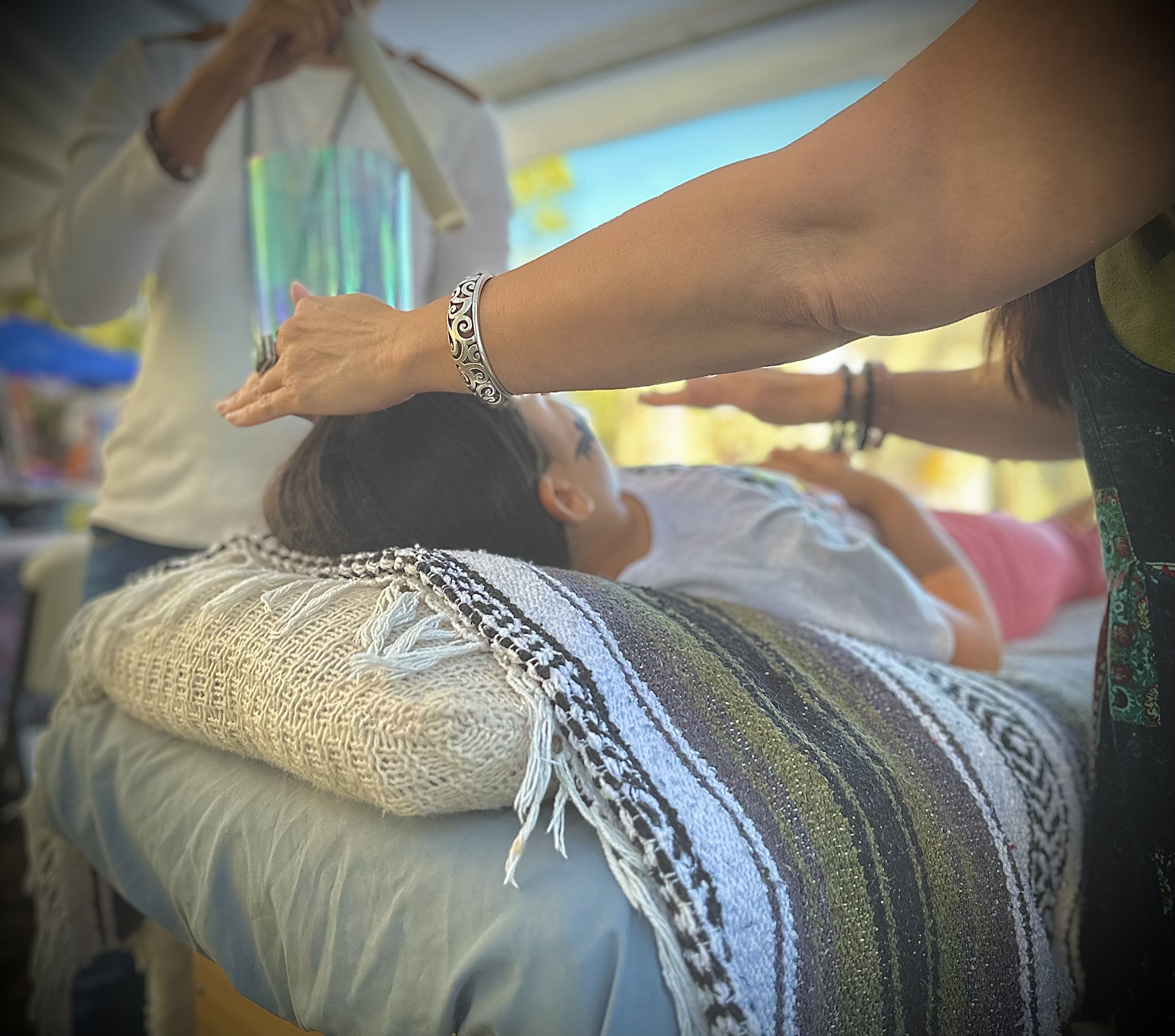 A person lying down on a colorful woven blanket, with their arm outstretched and wearing a decorative silver bracelet. The person's face is partially visible, and they appear to be outdoors in a sunny setting with other people in the background.
