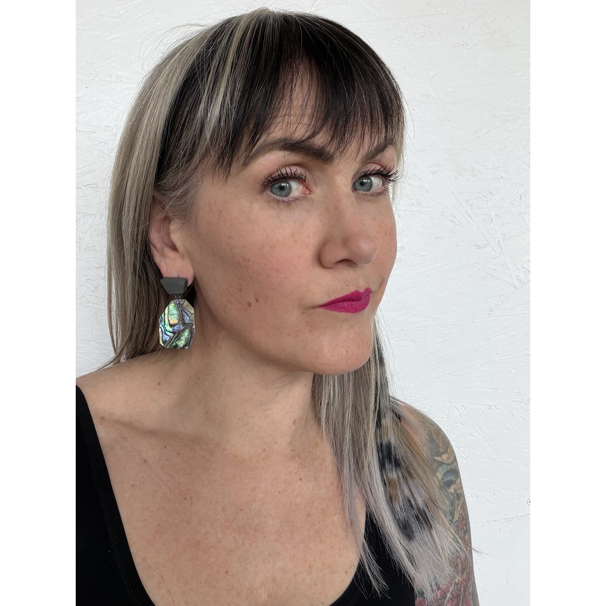 a woman with dark pink lipstick and blue eyes wearing irridescent green shell earrings and looking directly at the camera