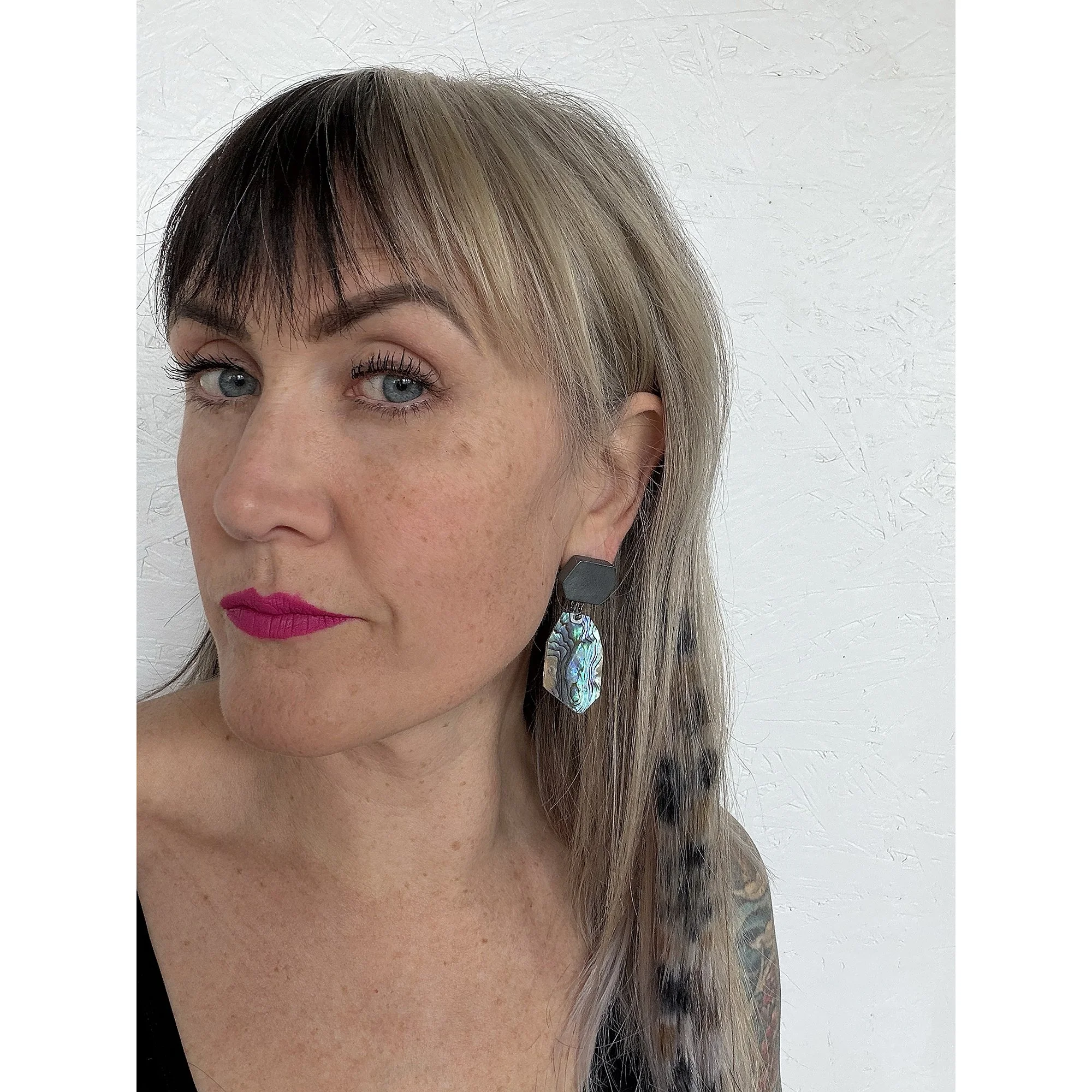 a woman with pink lipstick, big hair and bold eyebrows wearing dark green abalone shell earrings