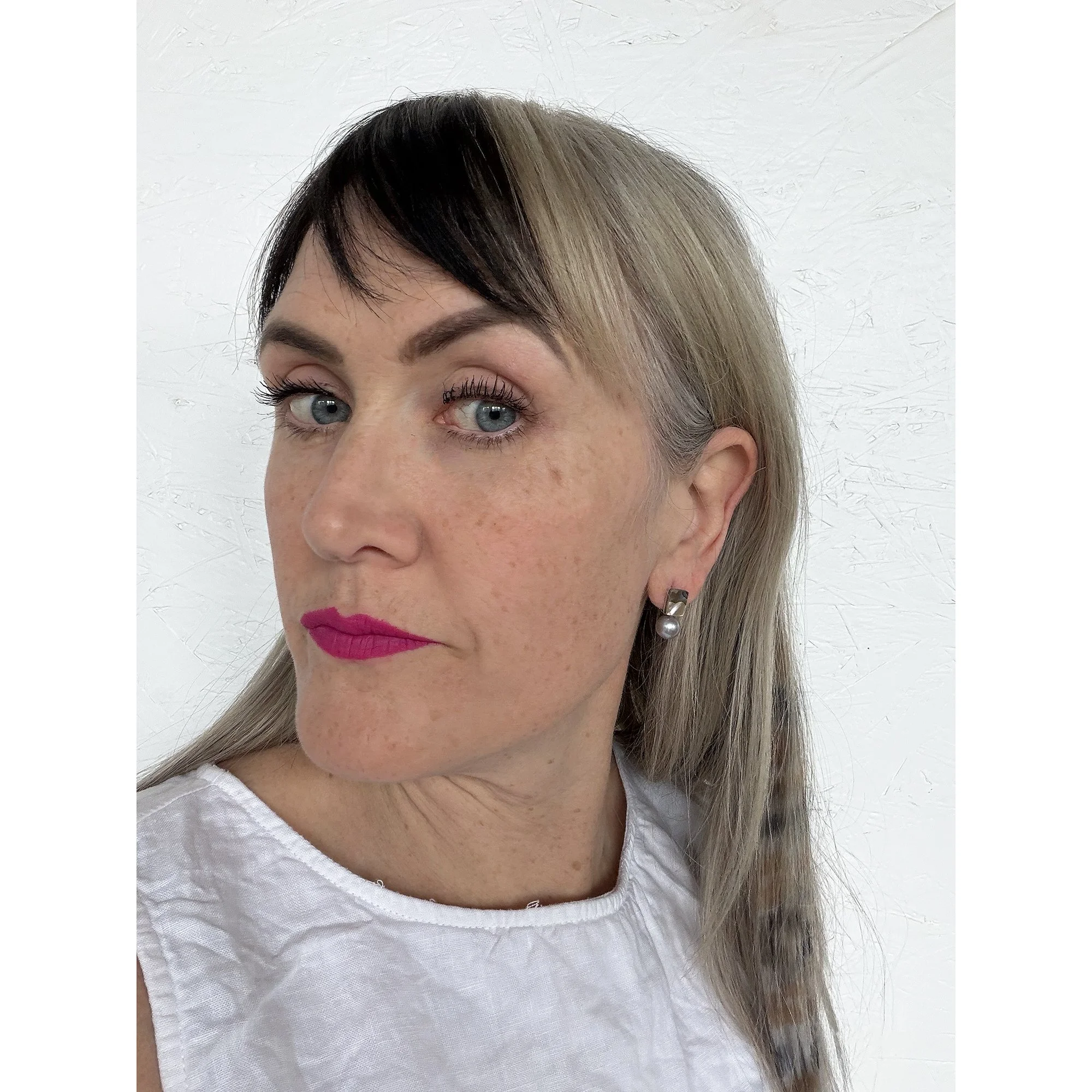 front view of woman with blonde hair, pink lipstick and white shirt wearing small pearl stud earrings with a rainbow of color on top and a round black pearl on the bottom