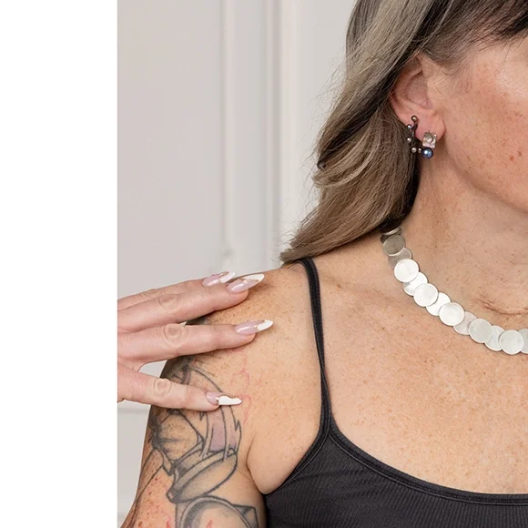 close up of small hoop earrings on woman with wavy hair shiny silver necklce wearing black tank top and tattoos