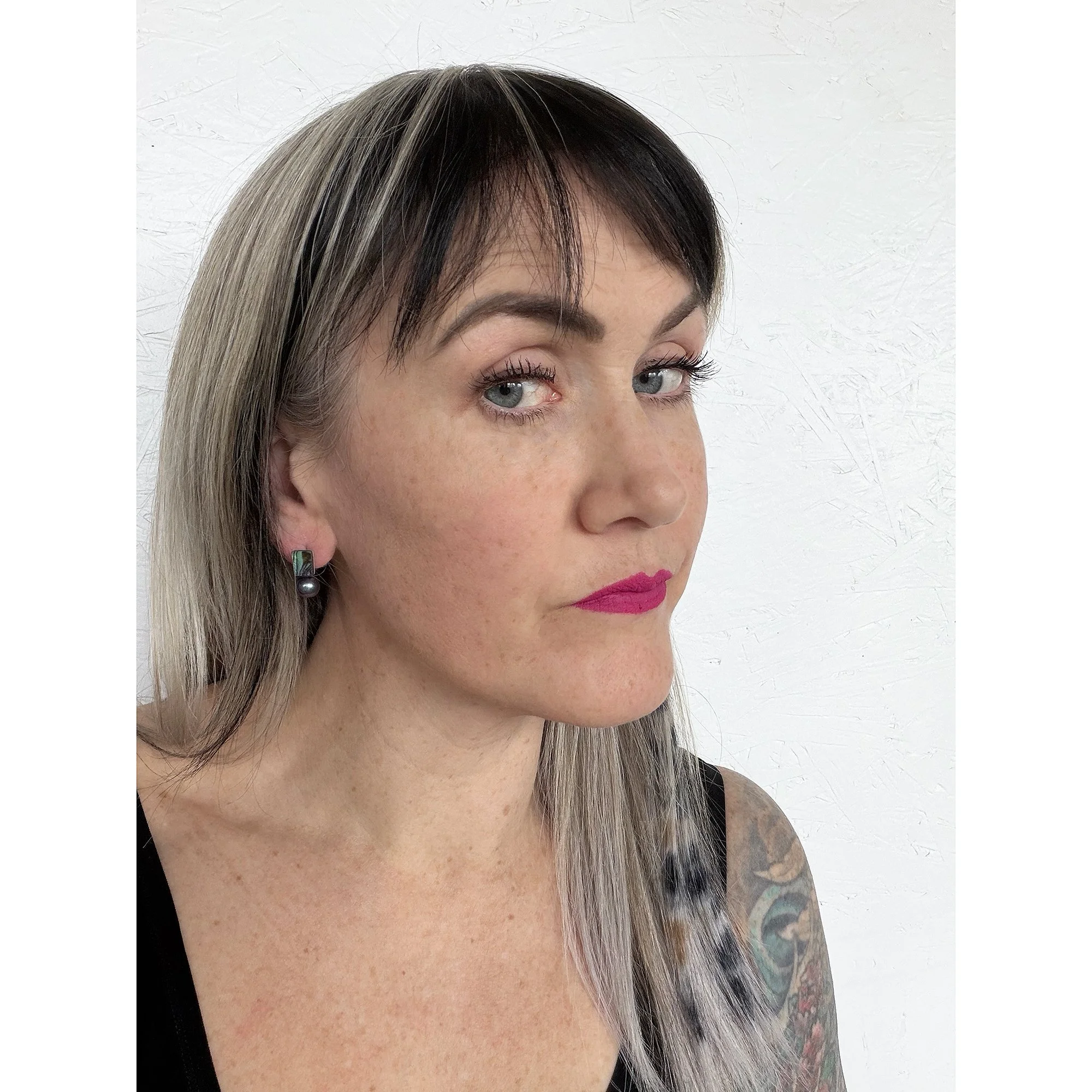 front view of woman with blonde hair, pink lipstick and black shirt wearing small dark green abalone and dark blue black peacock pearl earrings looking at camera