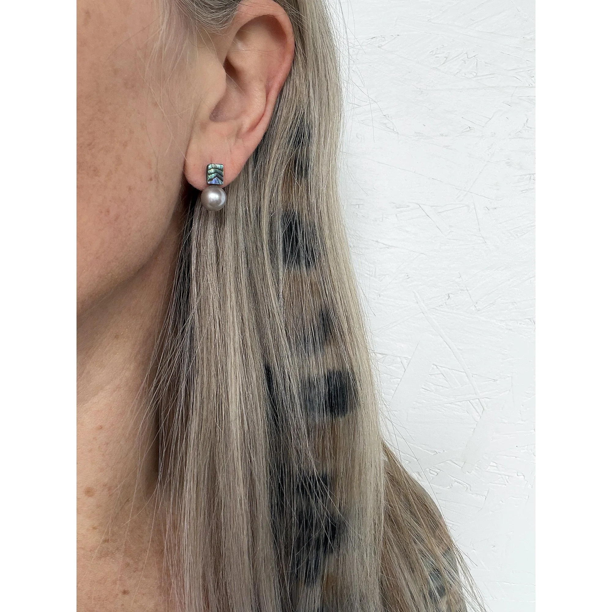 side view of woman with blonde and cheetah patterned hair and white shirt wearing small pearl stud earring with waves of color on top and a round silver pearl on the bottom
