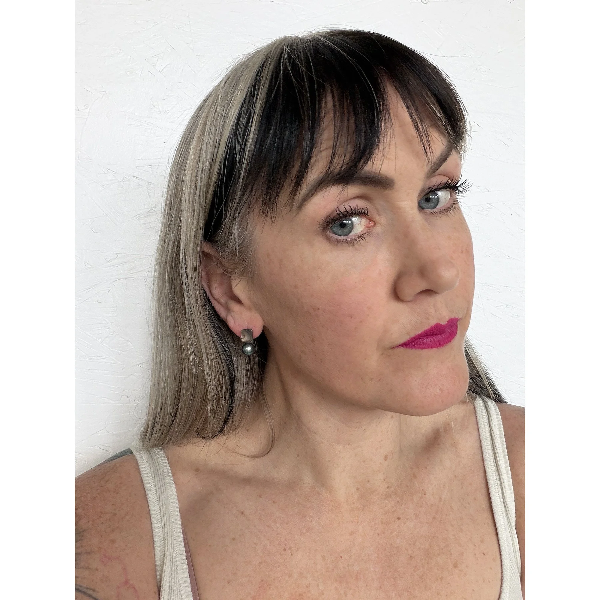 front view of woman with blonde hair, dark pink lipstick and white shirt wearing small pearl stud earring with a rainbow of color on top and a round black pearl on the bottom