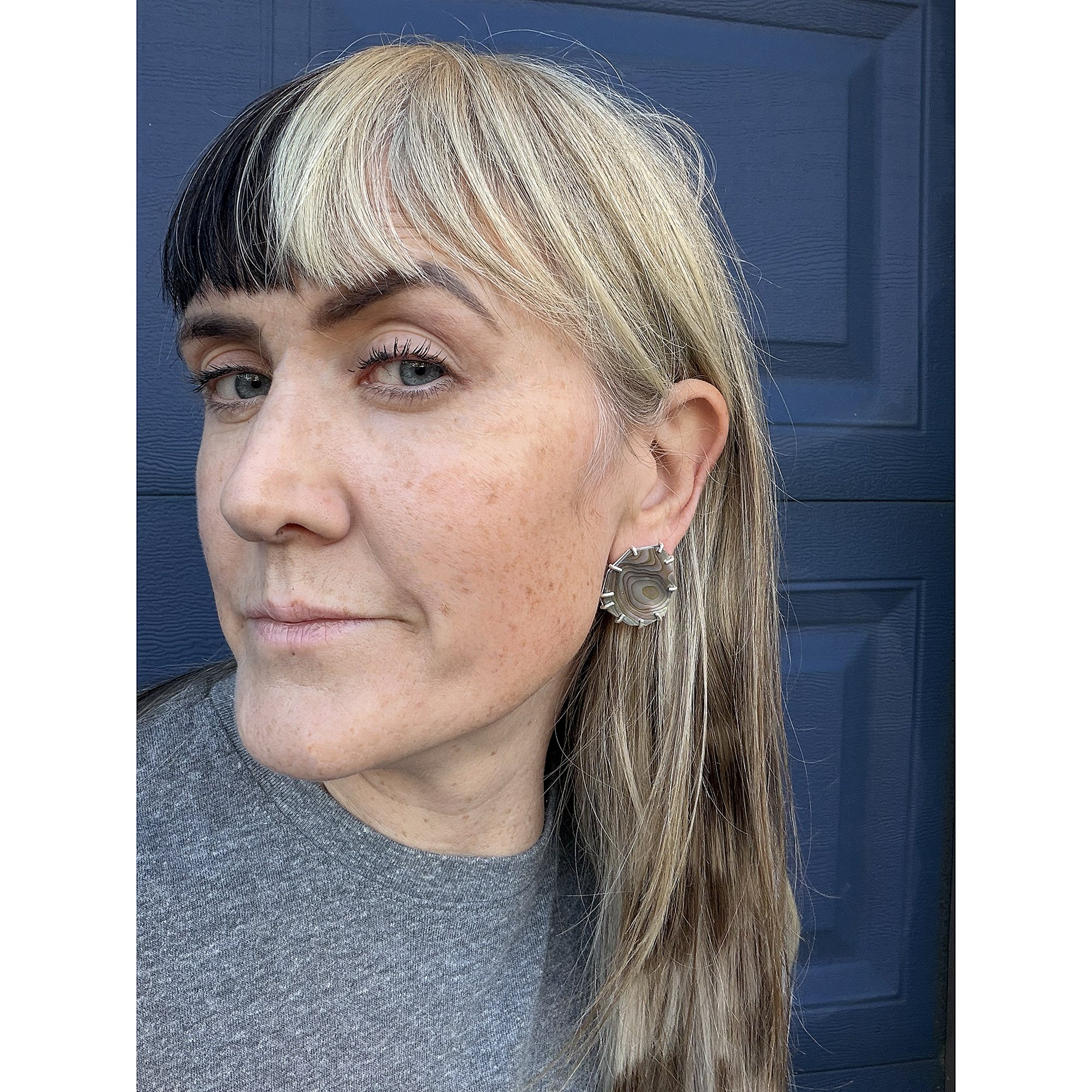 woman with white and black bangs wearing red abalone earrings in front of a blue background