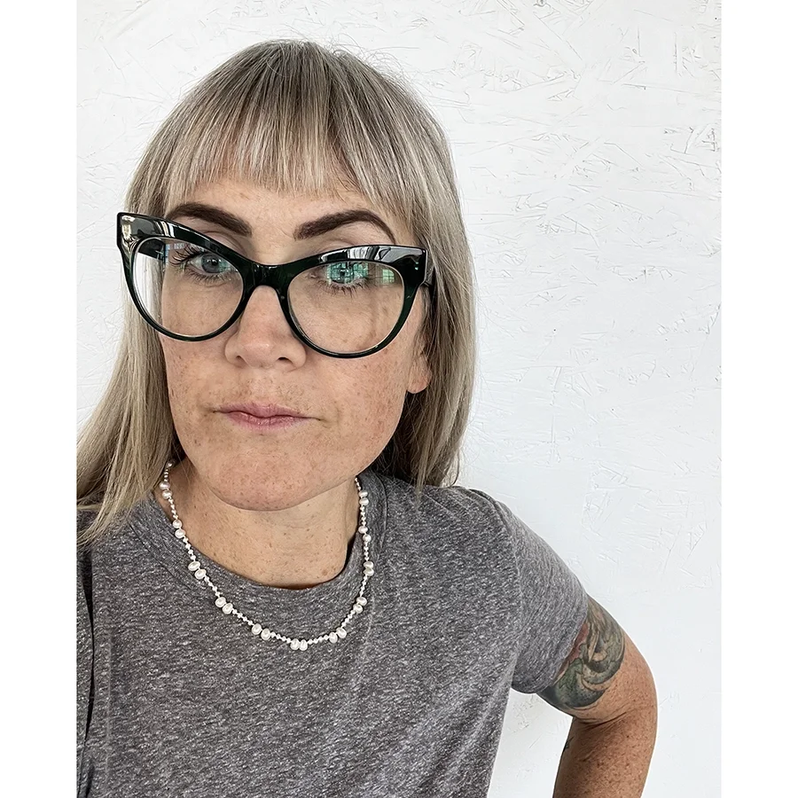 woman with blond hair and green glasses wearing gray tee shirt and delicate white pearl neckalace with alternating big and small pearls