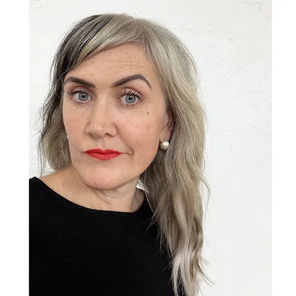 woman with black shirt blond wavy hair and red lips wearing small silver ball earrings with yellow gold lines
