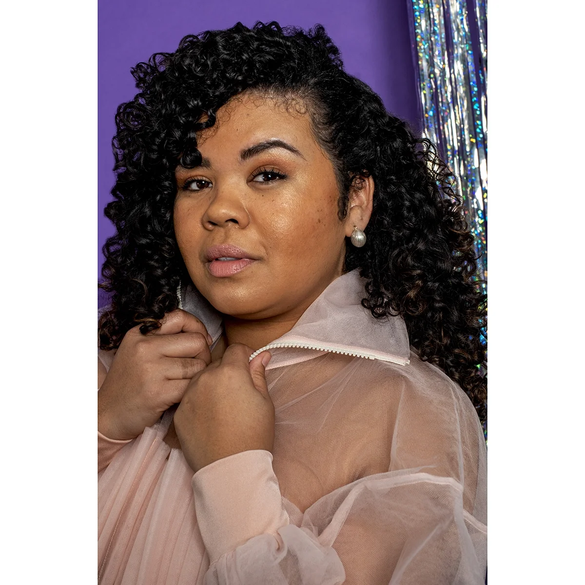 woman with dark curly hair and pin sheer jacket wearing small shiny sterling silver earrings in front of purple and shimmery metallic wall
