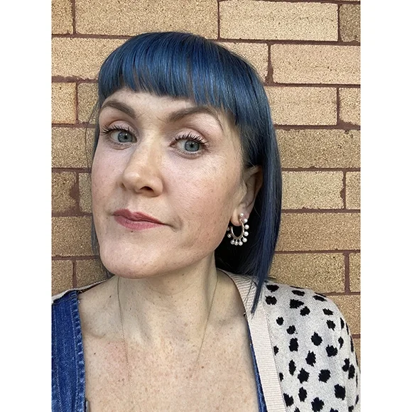 woman with blue hair facing camera wearing silver hoop earrings with white pearls in front of brick building