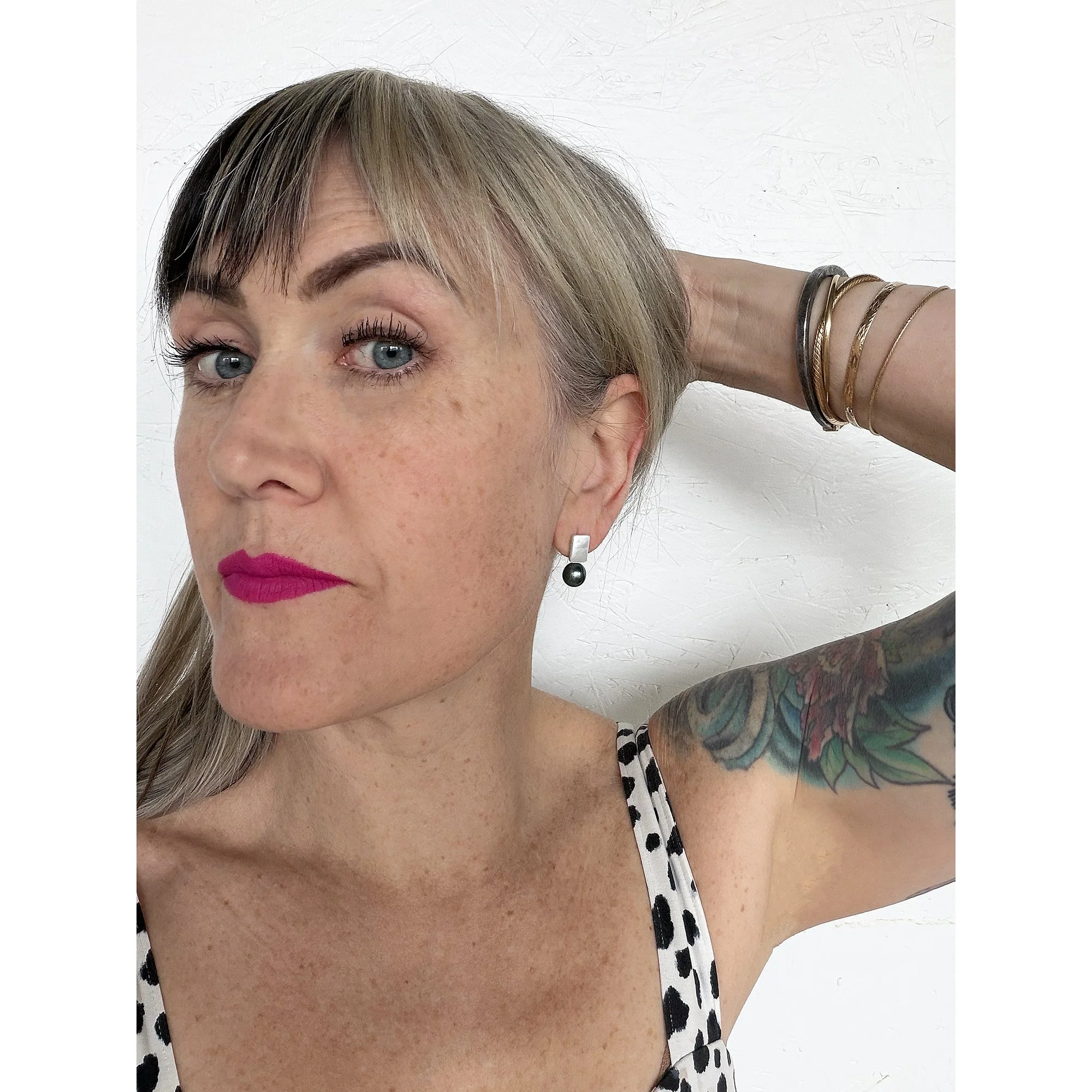 woman with pink lipstick wearing white and black cheetah shirt wearing small earrings with shimmery mother of pearl and a perfect big deep blue black and green pearl