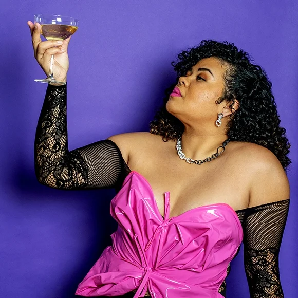 beatiful brown woman with black curly hair wearing black and white pearl earrings holding a glass of champagne in a pink latex bow top in front of a purple and white backdrop
