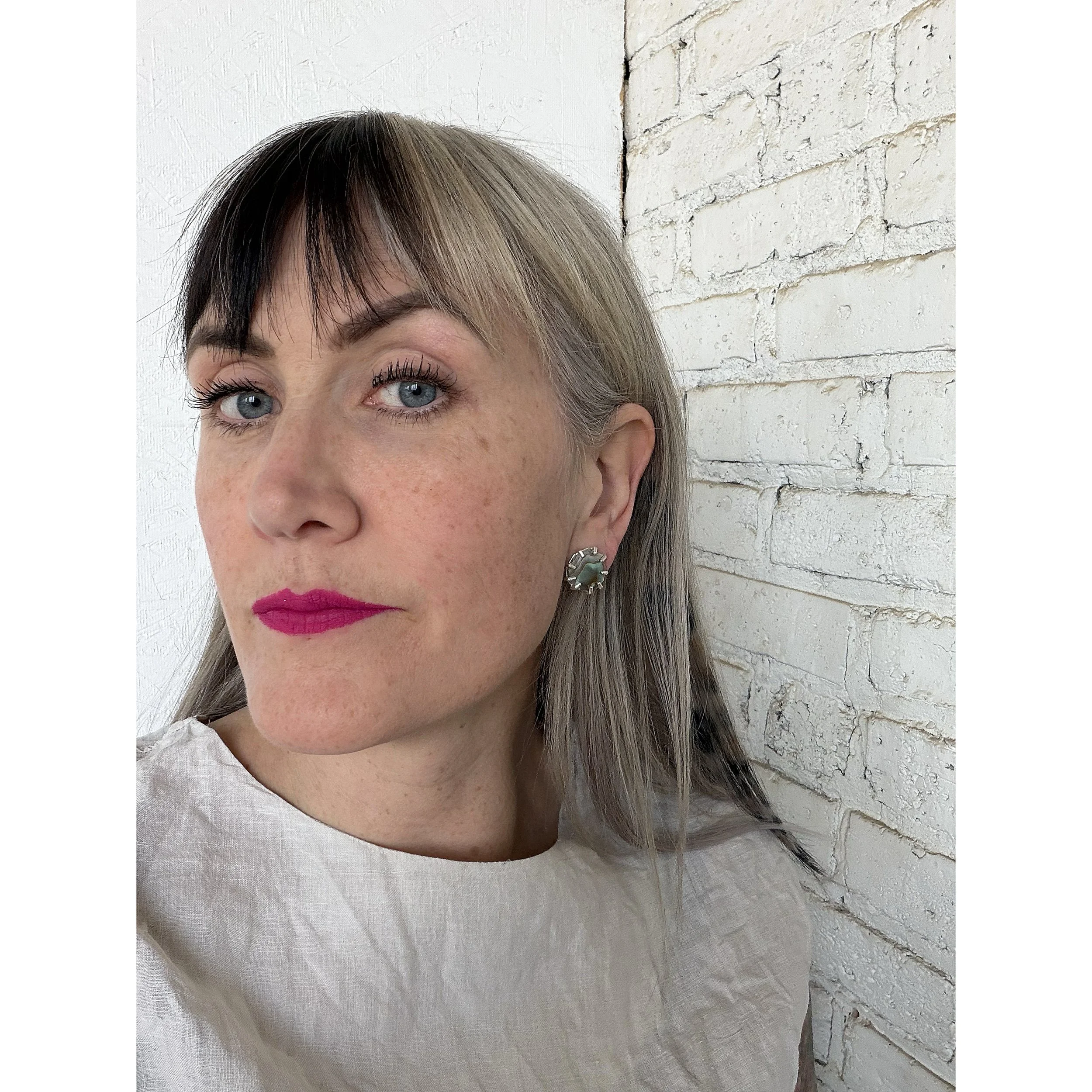 blonde woman's ear with shimmery small red and pink abalone post and silver prongs with white brick background