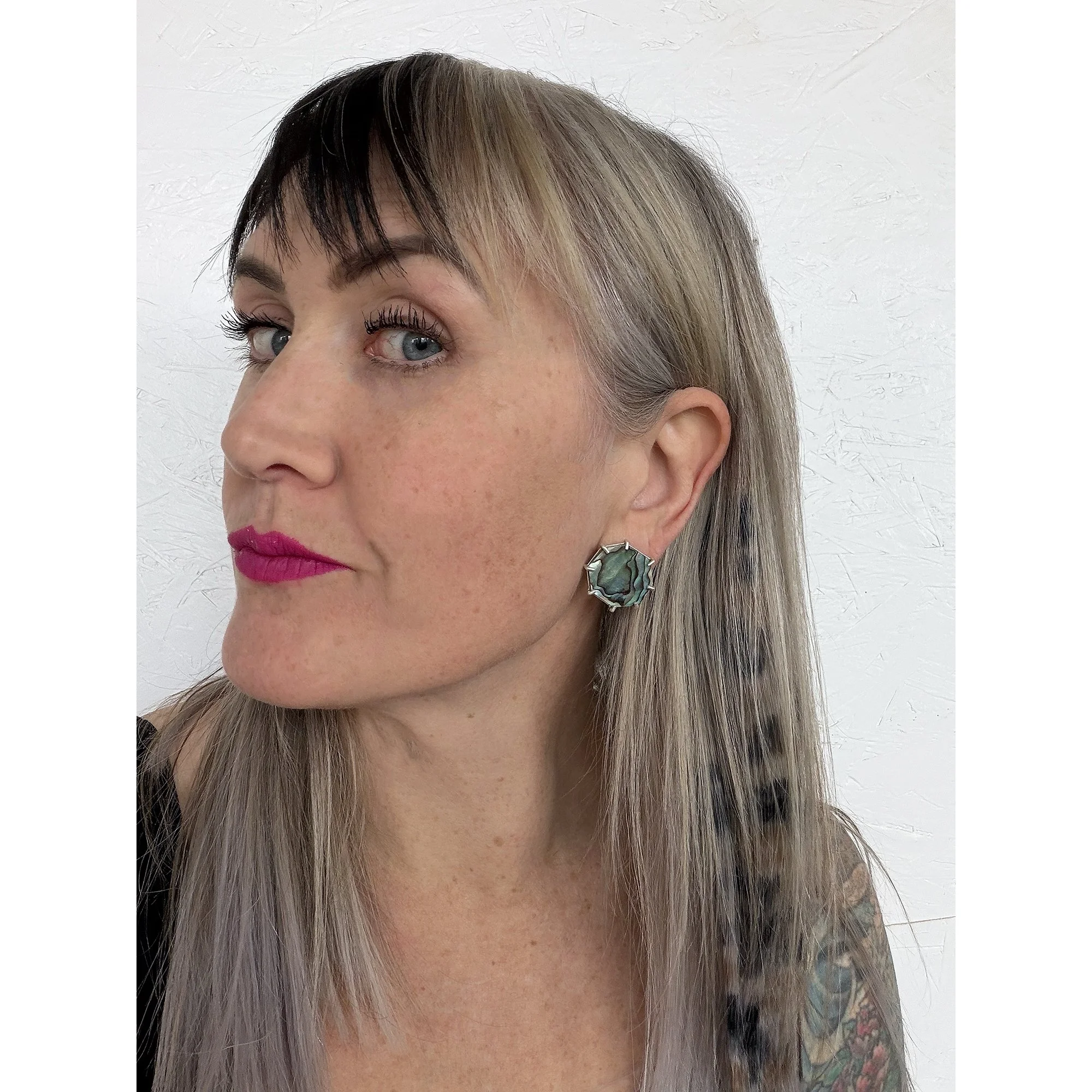 a woman with blue eyes and pink lipstick wearing dark green shiny abalone post earrings looking at the camera
