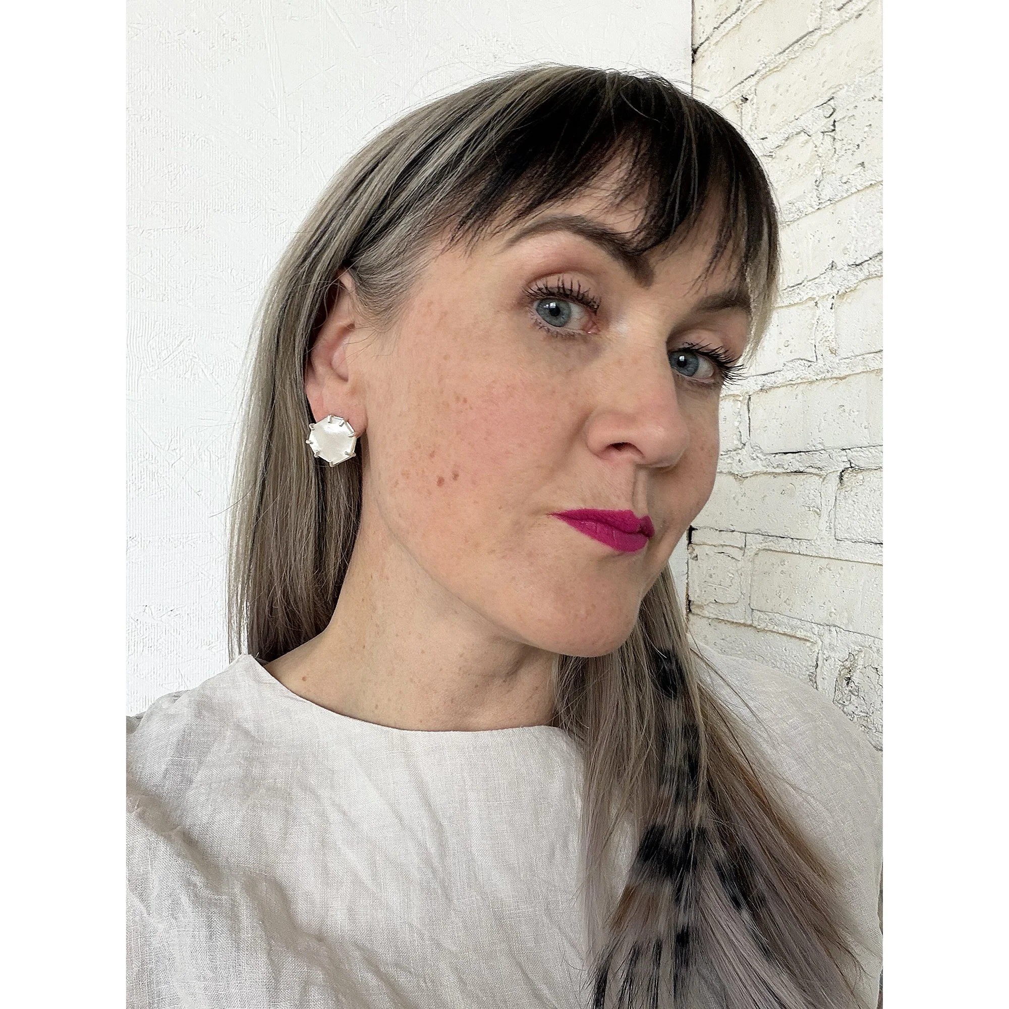 woman with blonde hair wearing mother of pearl post earrings in room with white brick