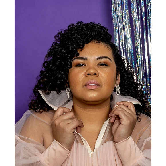 small pearl earrings on woman with dark curly hair pink lips and soft pink jacket with a purple and silver glitter background