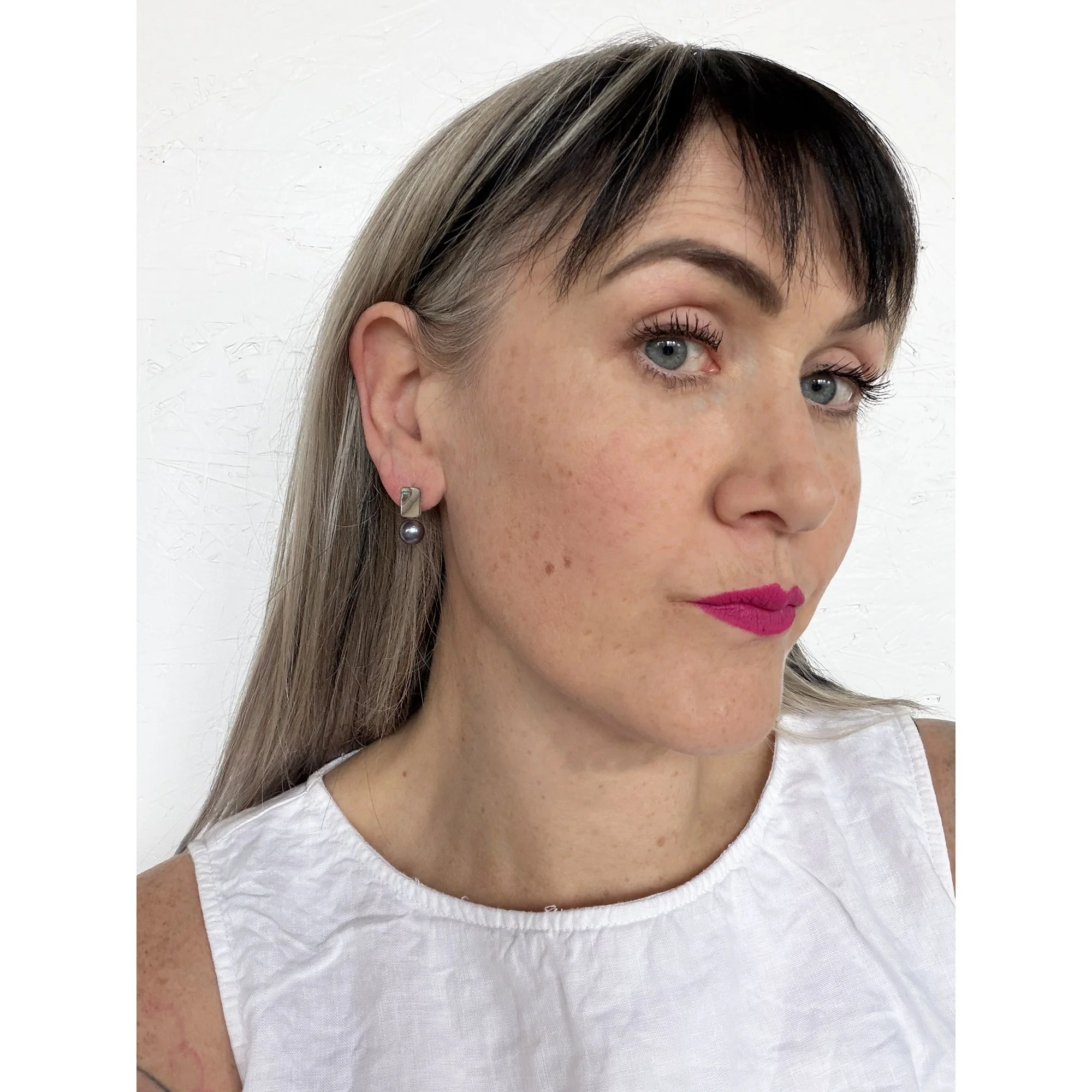 front view of woman with blonde hair, pink lipstick and white shirt wearing small pearl stud earring with a rainbow of color on top and a round black pearl on the bottom